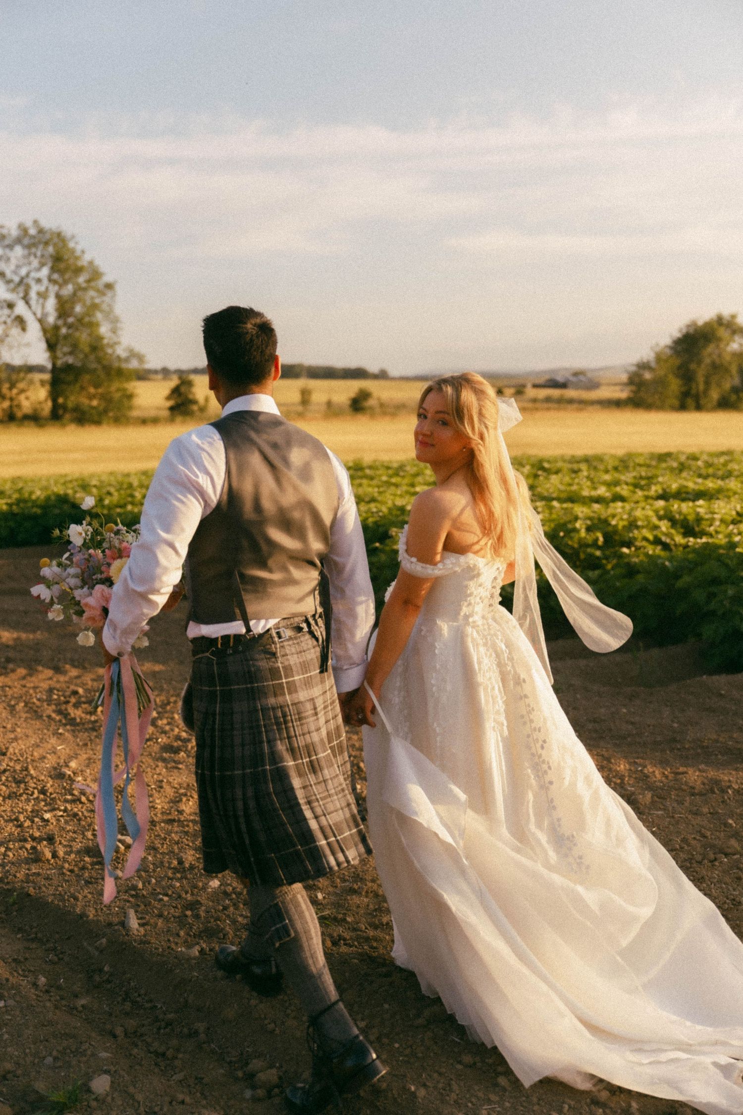 A couple stands in a golden field at sunset as her flowing white wedding dress and veil catches the evening breeze.