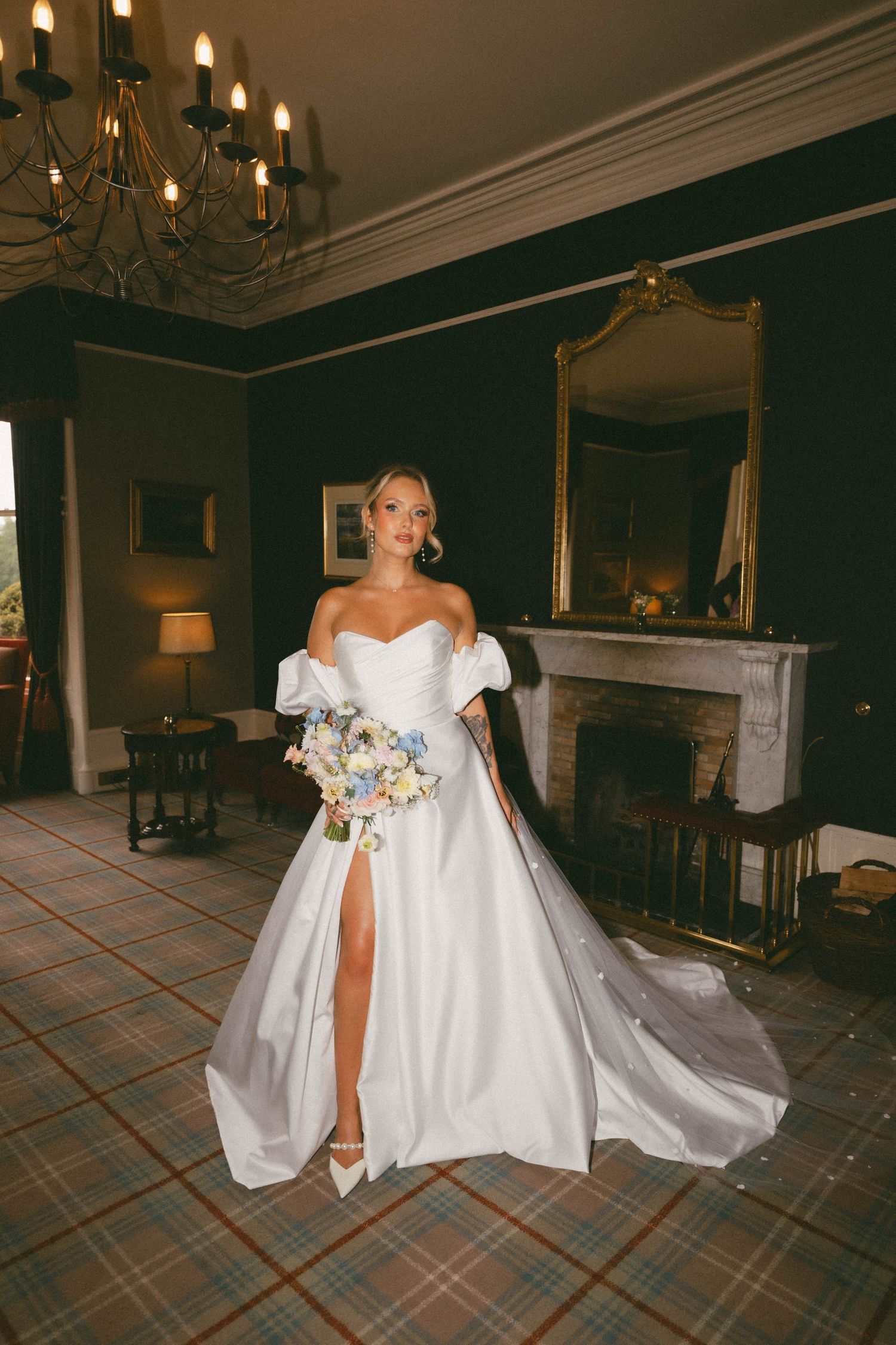 Elegant bride in a flowing white ball gown poses against plaid carpet in an ornate room with chandeliers.