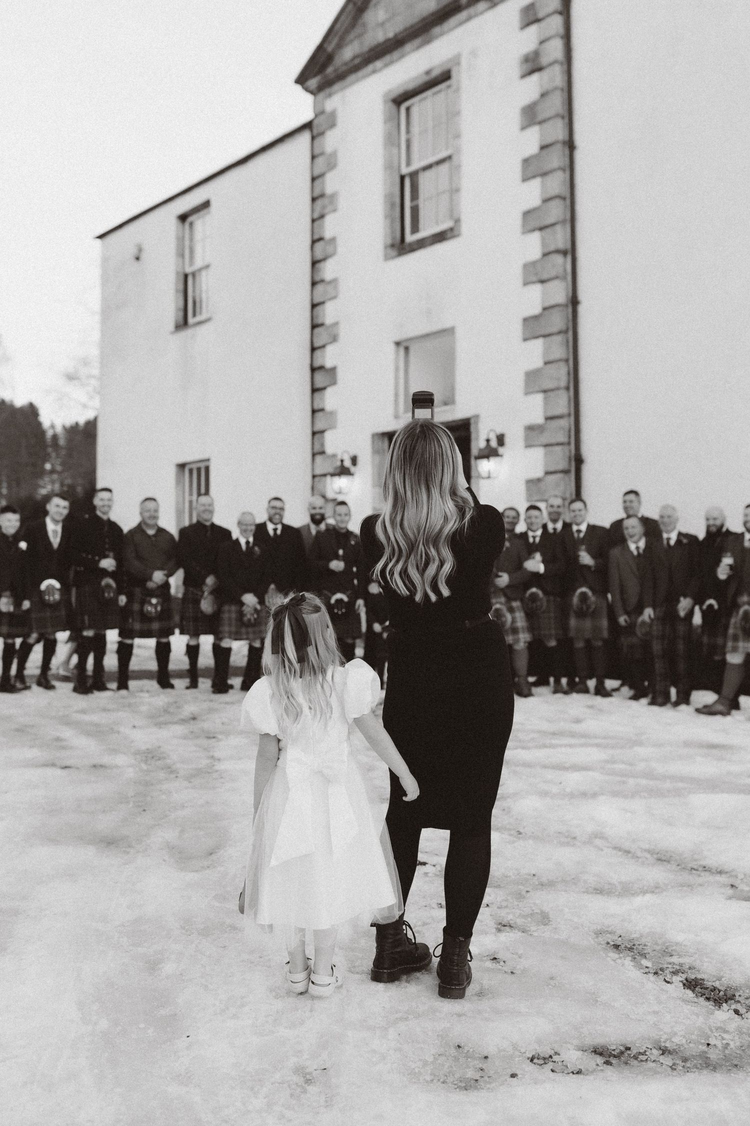 Black and white wedding photo showing a woman in black and child in white dress facing a group of kilted men at a venue.