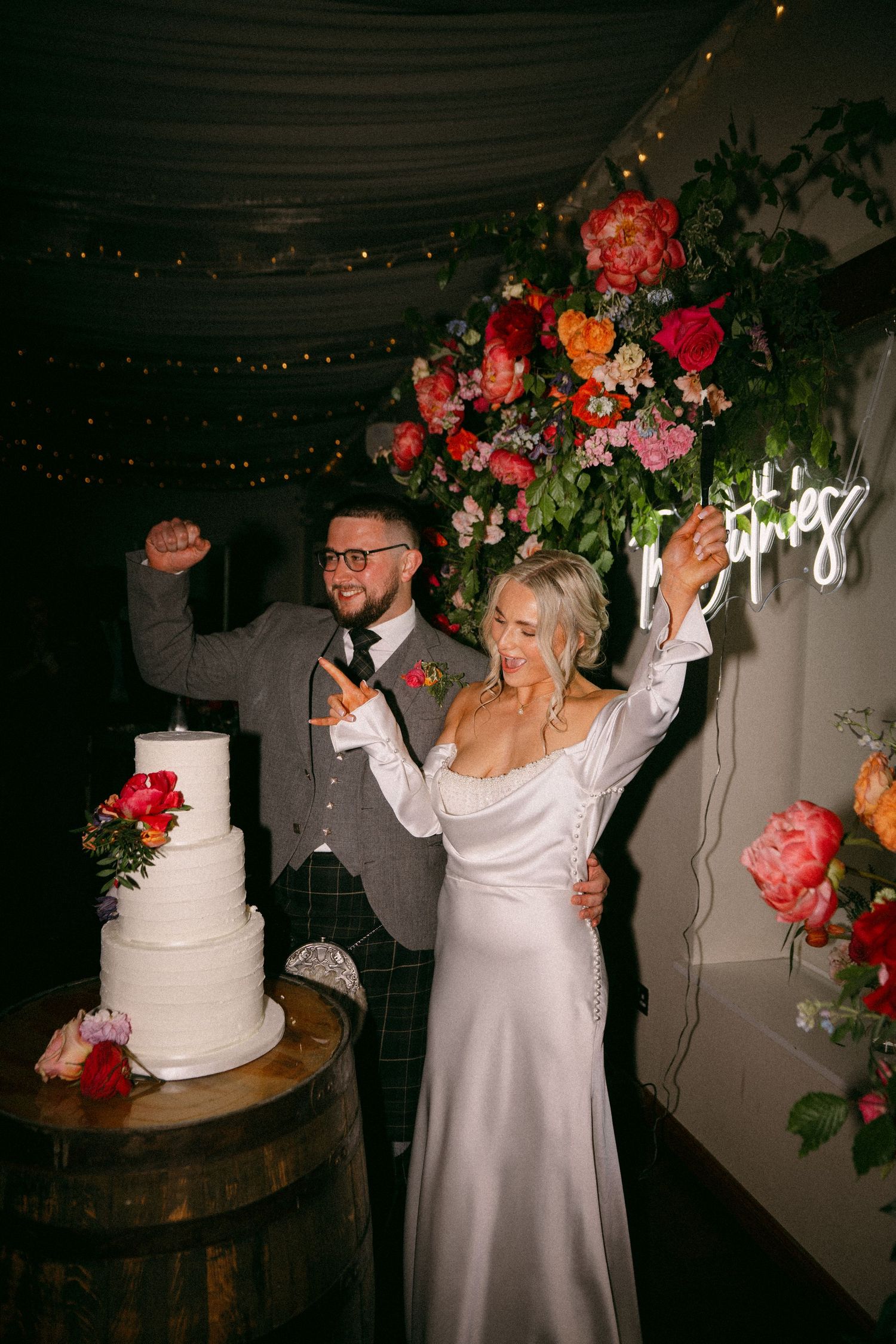 Newlyweds celebrate joyfully next to their wedding cake surrounded by floral decorations. Aberdeen wedding photographer, Logie country house