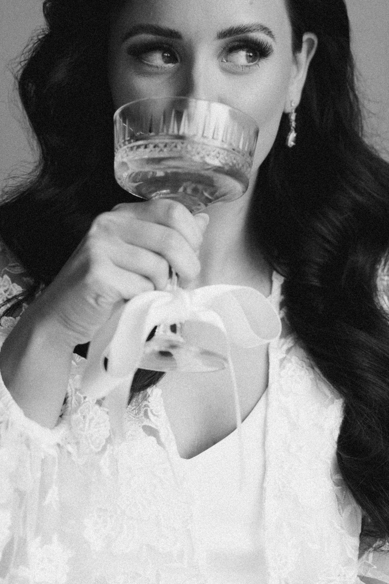A black and white photo of someone in white lace sipping from an ornate crystal champagne glass.
