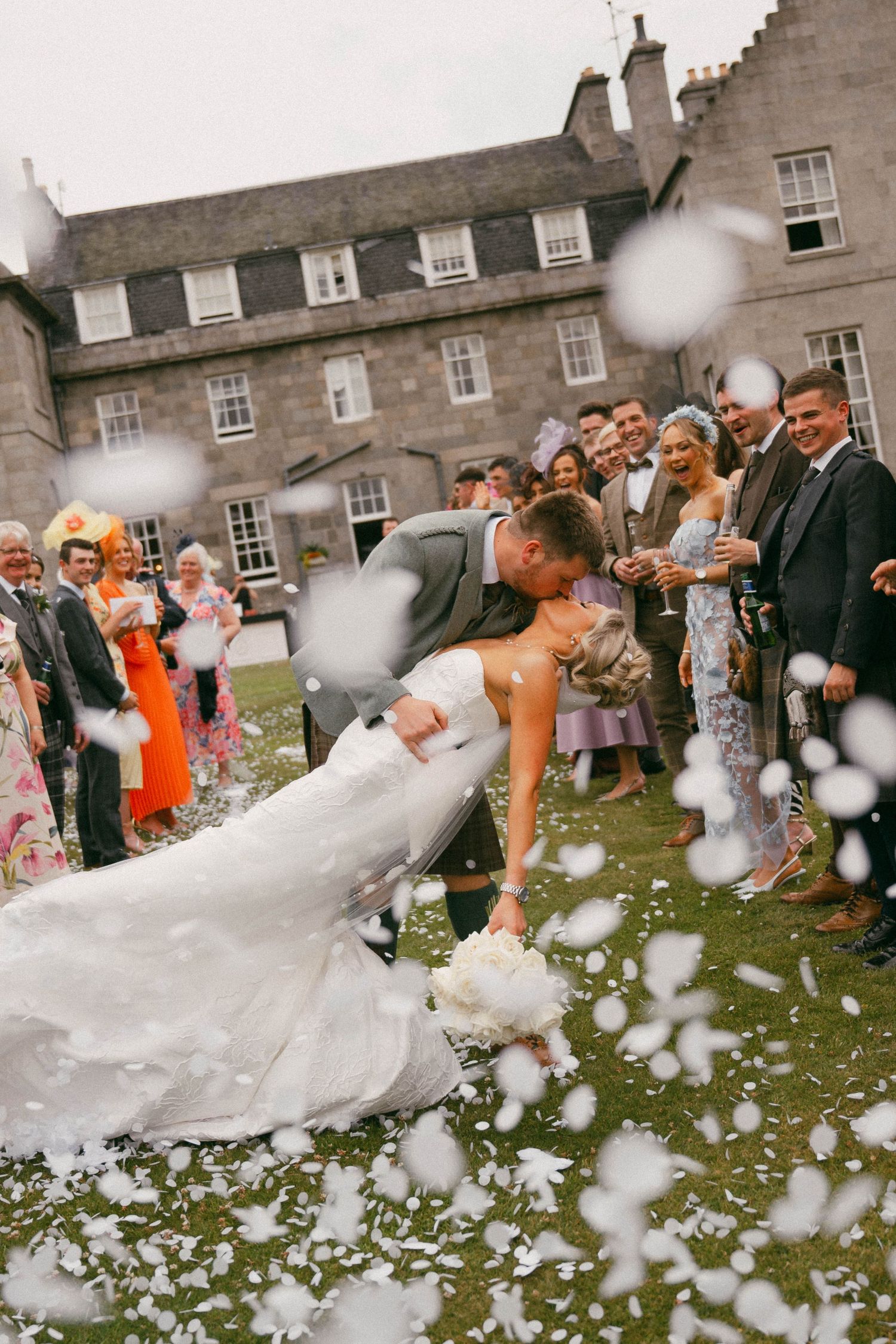 Wedding guests gather outdoors next to historic stone building as white confetti fills the air during celebration.