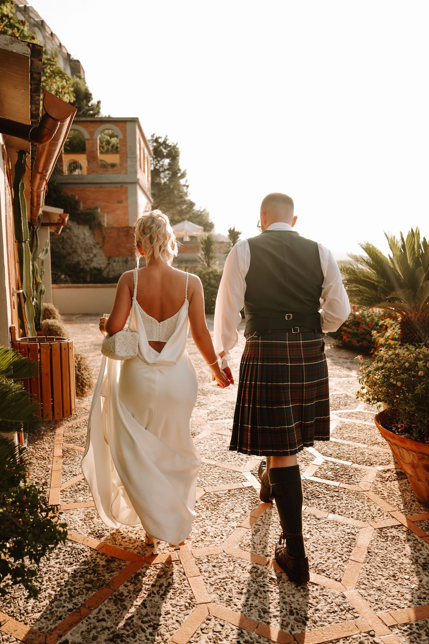 Couple walking along a sunny Mediterranean pathway with groom wearing traditional Scottish kilt at sunset.