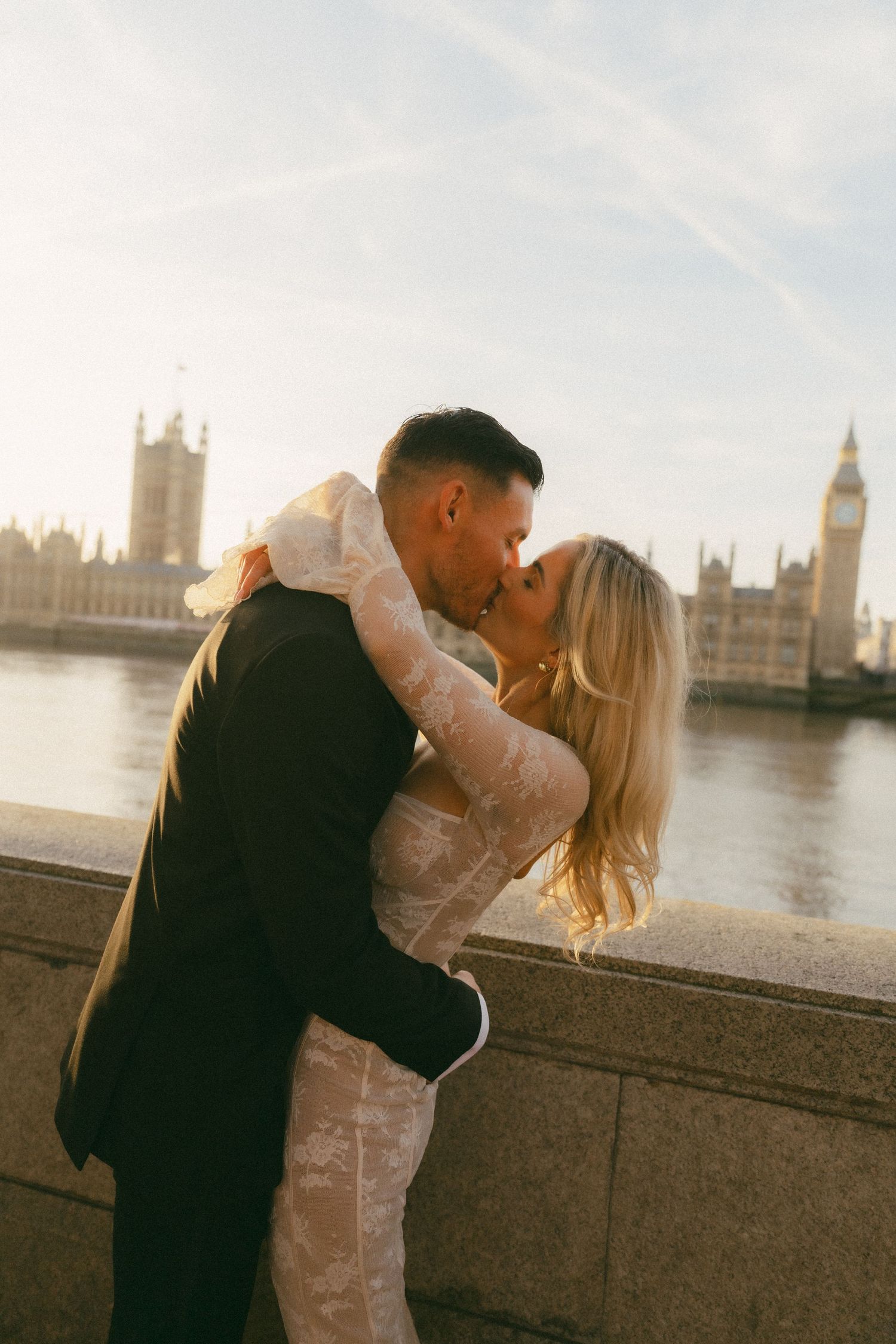 A romantic couple shares a kiss along the Thames River with London's Houses of Parliament and Big Ben in the background.