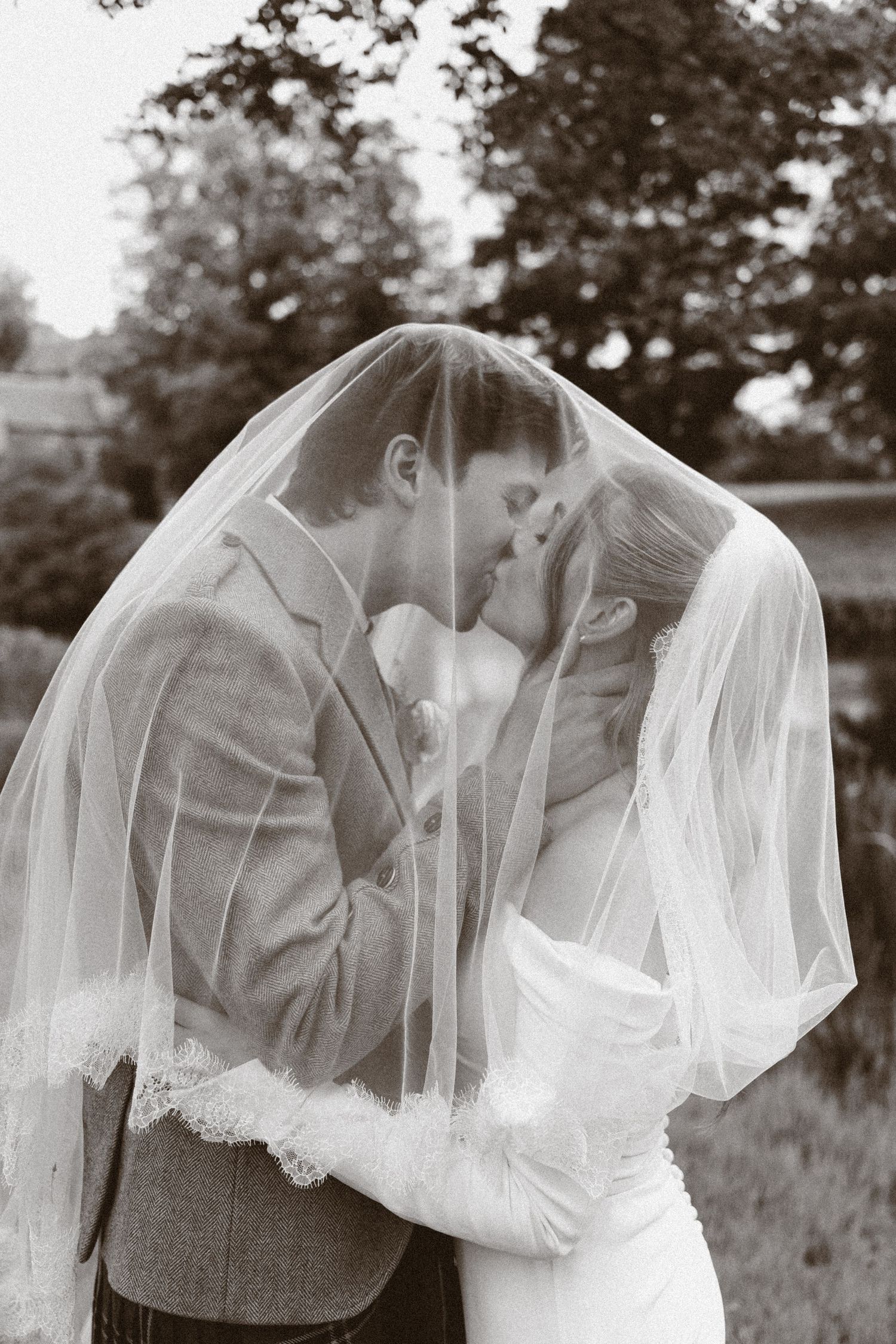 Black and white photo of bride and groom kissing under wedding veil outdoors.