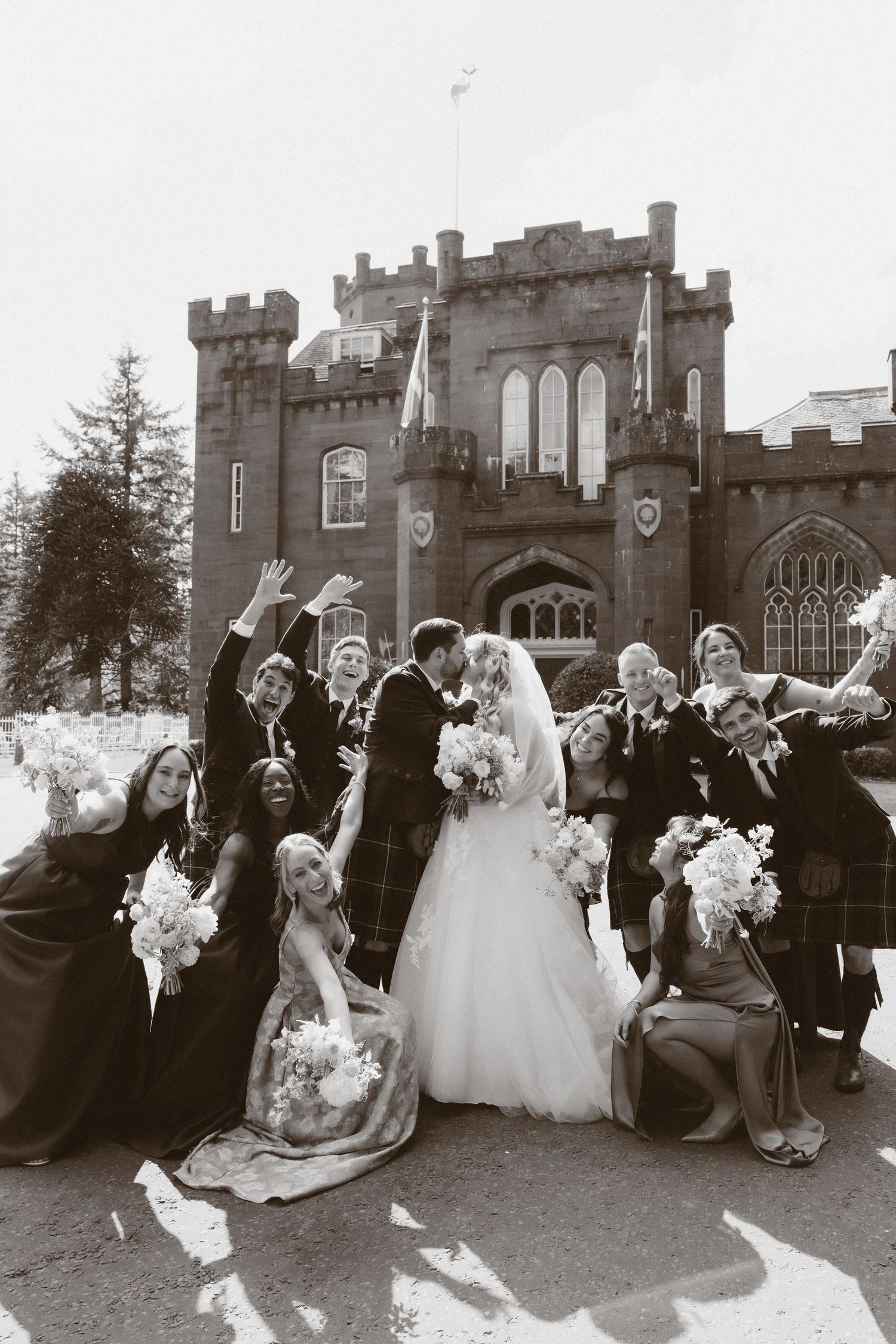 Wedding party celebrating outside a Scottish castle,bride and groom kissing while bridesmaids and groomsmen in kilts cheer.