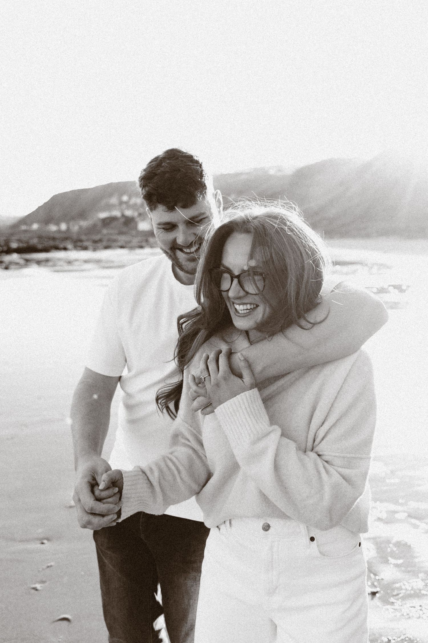 Black and white photo of a happy couple embracing and laughing together on a sandy beach with mountains behind.