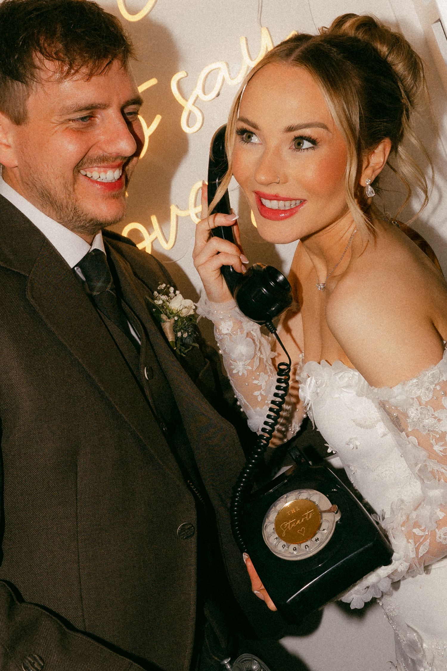 Bride in white lace gown holds vintage black telephone while groom in suit smiles beside her at wedding reception.