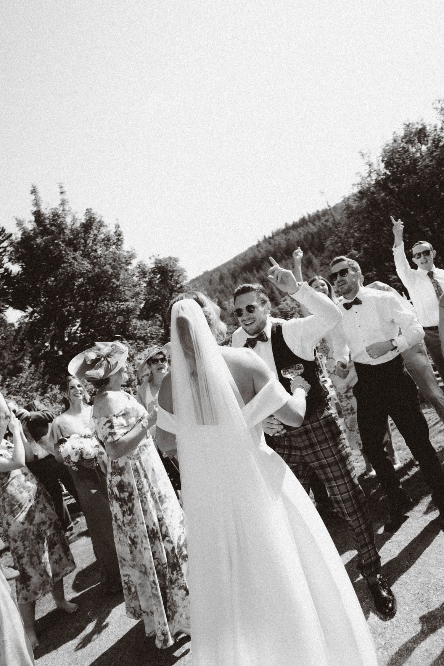 A black and white photograph of a festive outdoor wedding celebration with guests celebrating on a sunny day.