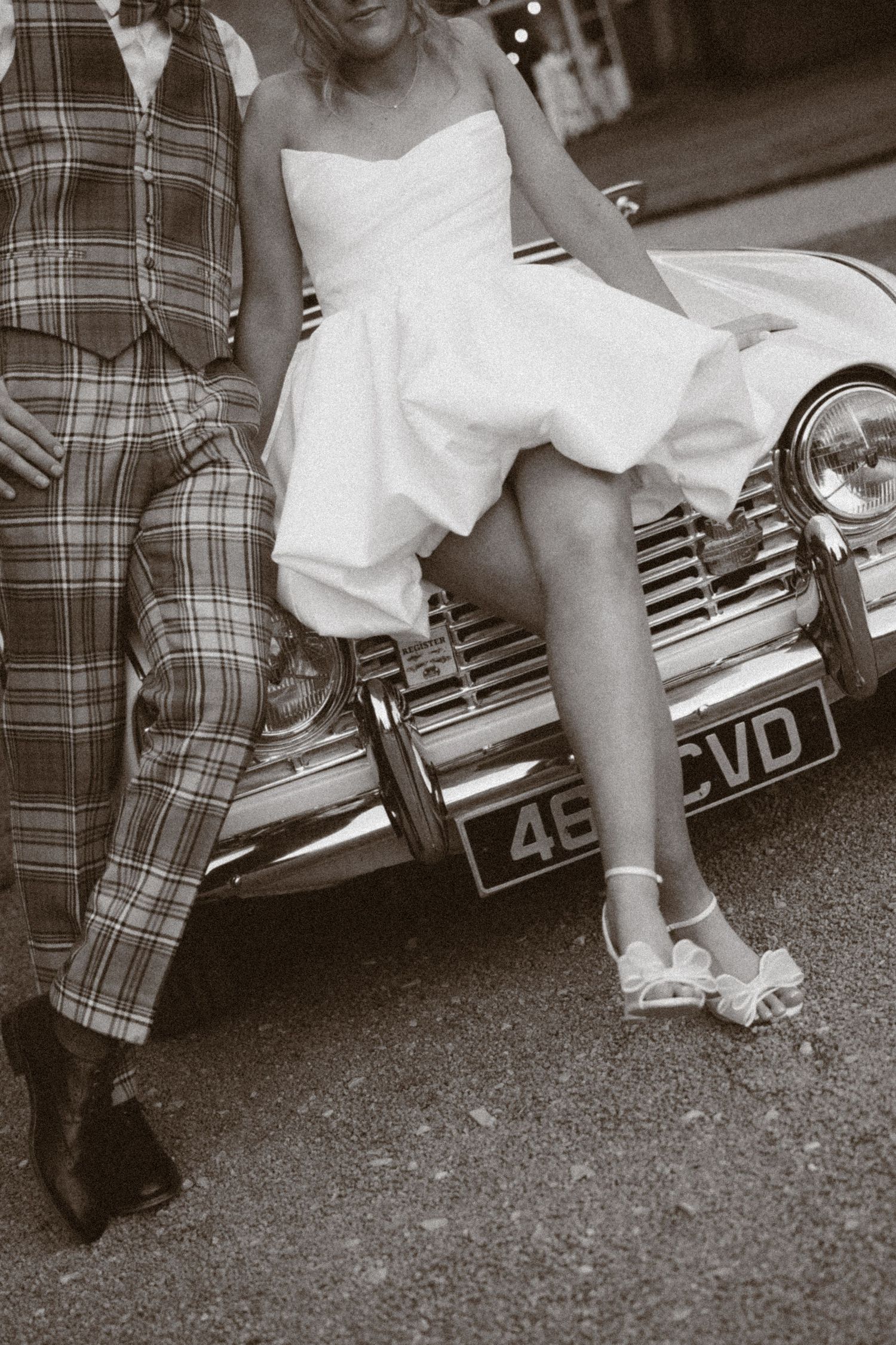 Bride in white dress and groom in plaid suit posing against a vintage classic car bumper on their wedding day.