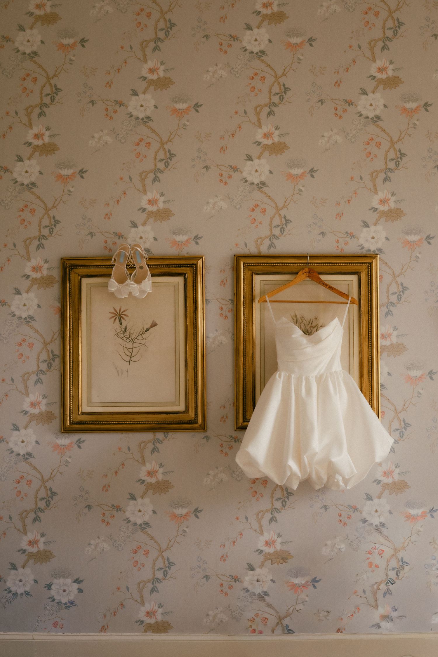 Short white wedding dress on gold hanger beside framed botanical art and heels on floral wallpaper.