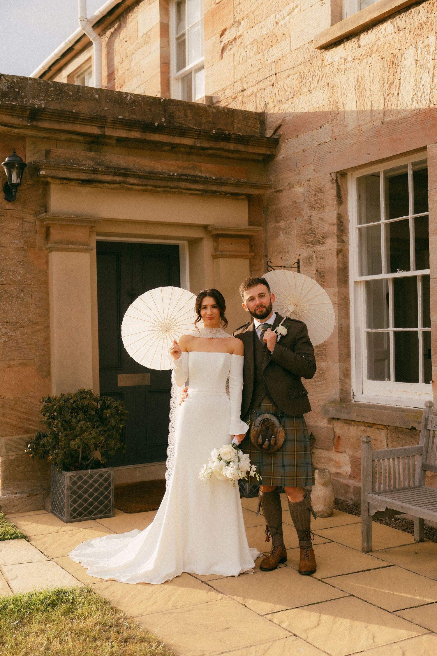 Bride and groom posing with white parasols outside a Scottish stone manor, groom wearing a tartan kilt.