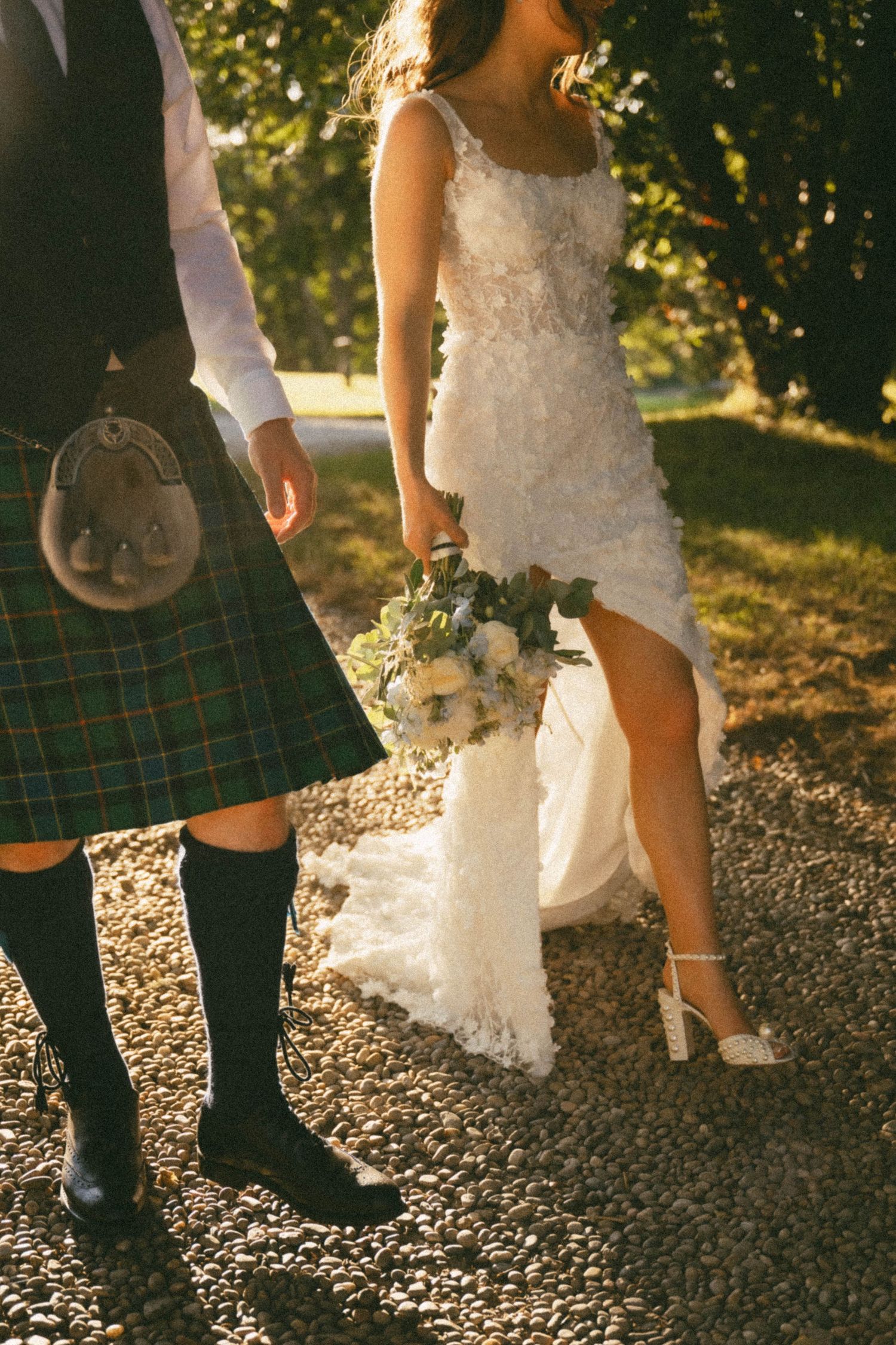 A bride in white lace dress holds a bouquet next to someone wearing a traditional Scottish kilt in outdoor sunlit setting.