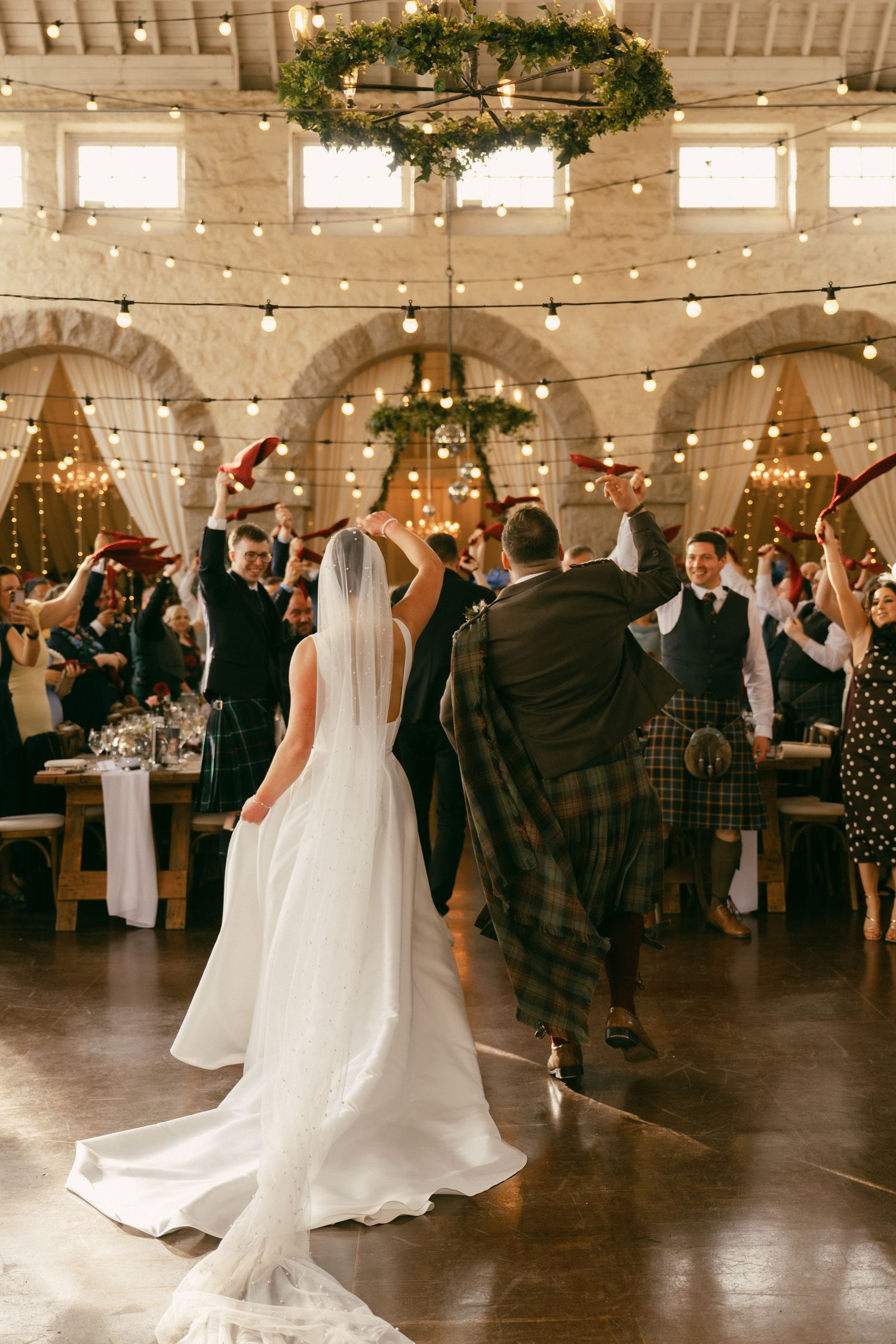 Bride and groom enter wedding reception hall with cheering guests, string lights, and greenery chandelier decor.