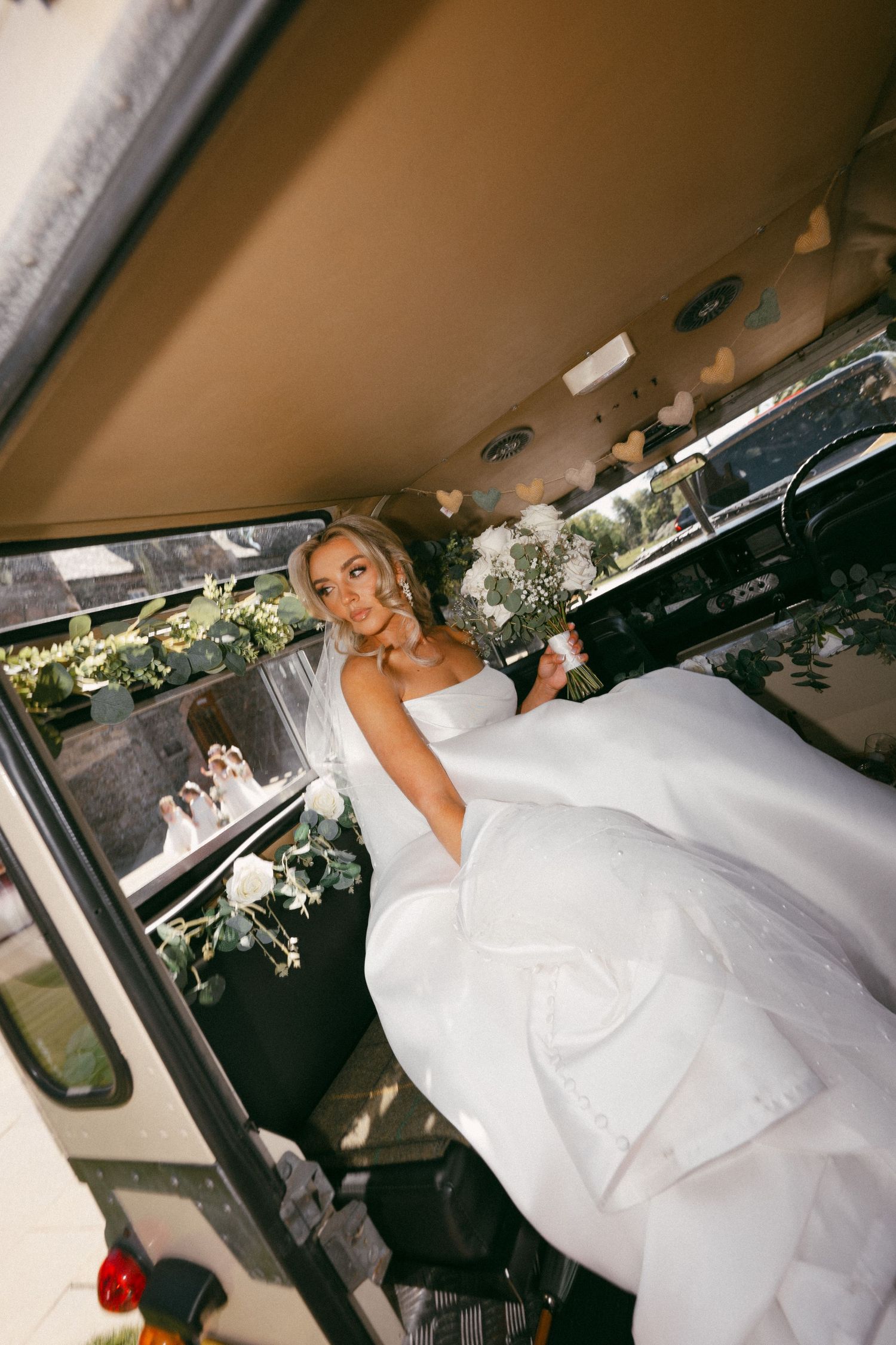 A bride in a stunning white ballgown wedding dress sits elegantly in the back of a decorated limo.