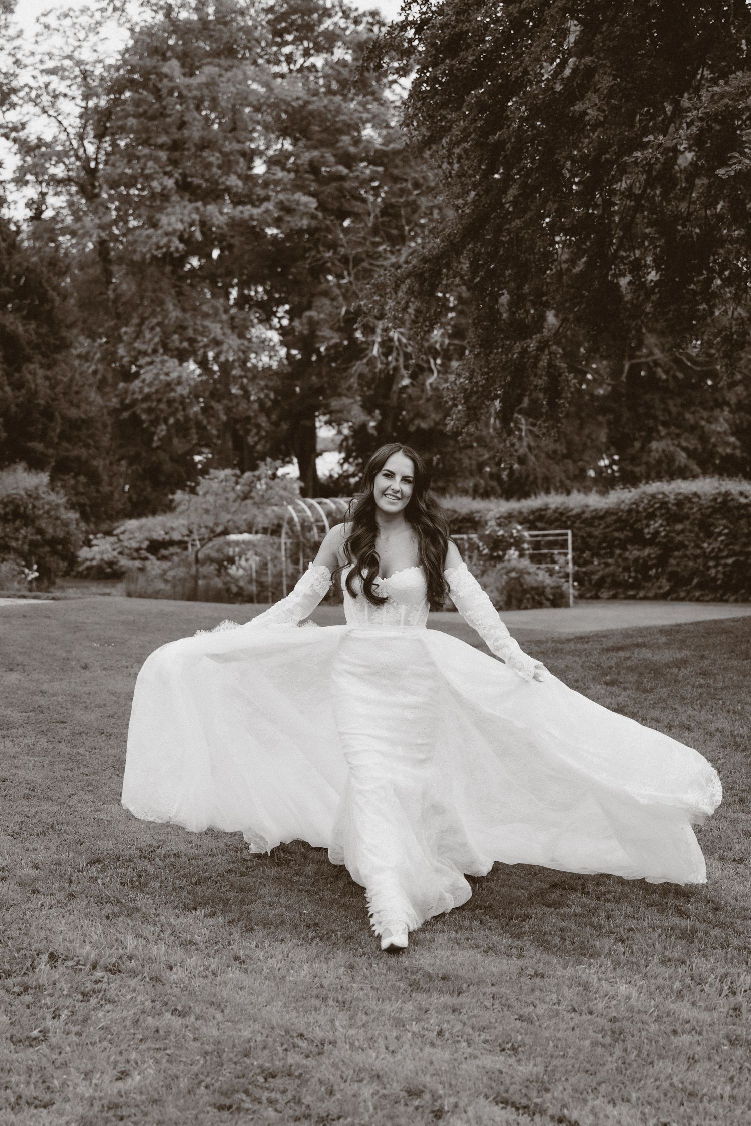 Black and white photo of a bride in a flowing wedding gown twirling outdoors in a lush garden setting.