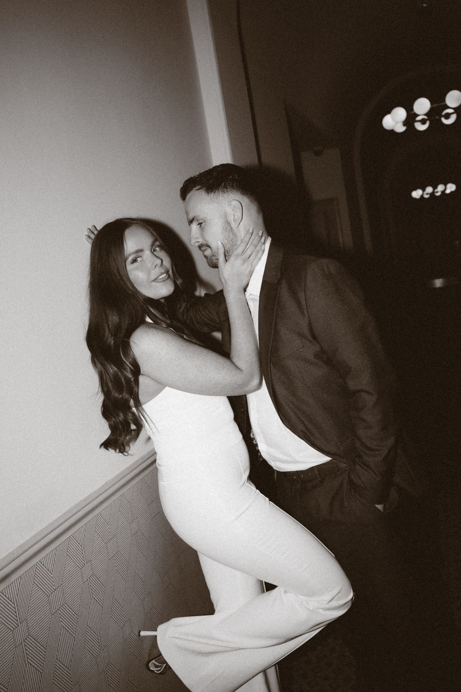 Black and white photo of a couple in formal wear, woman in white jumpsuit embracing a man in a dark suit.
