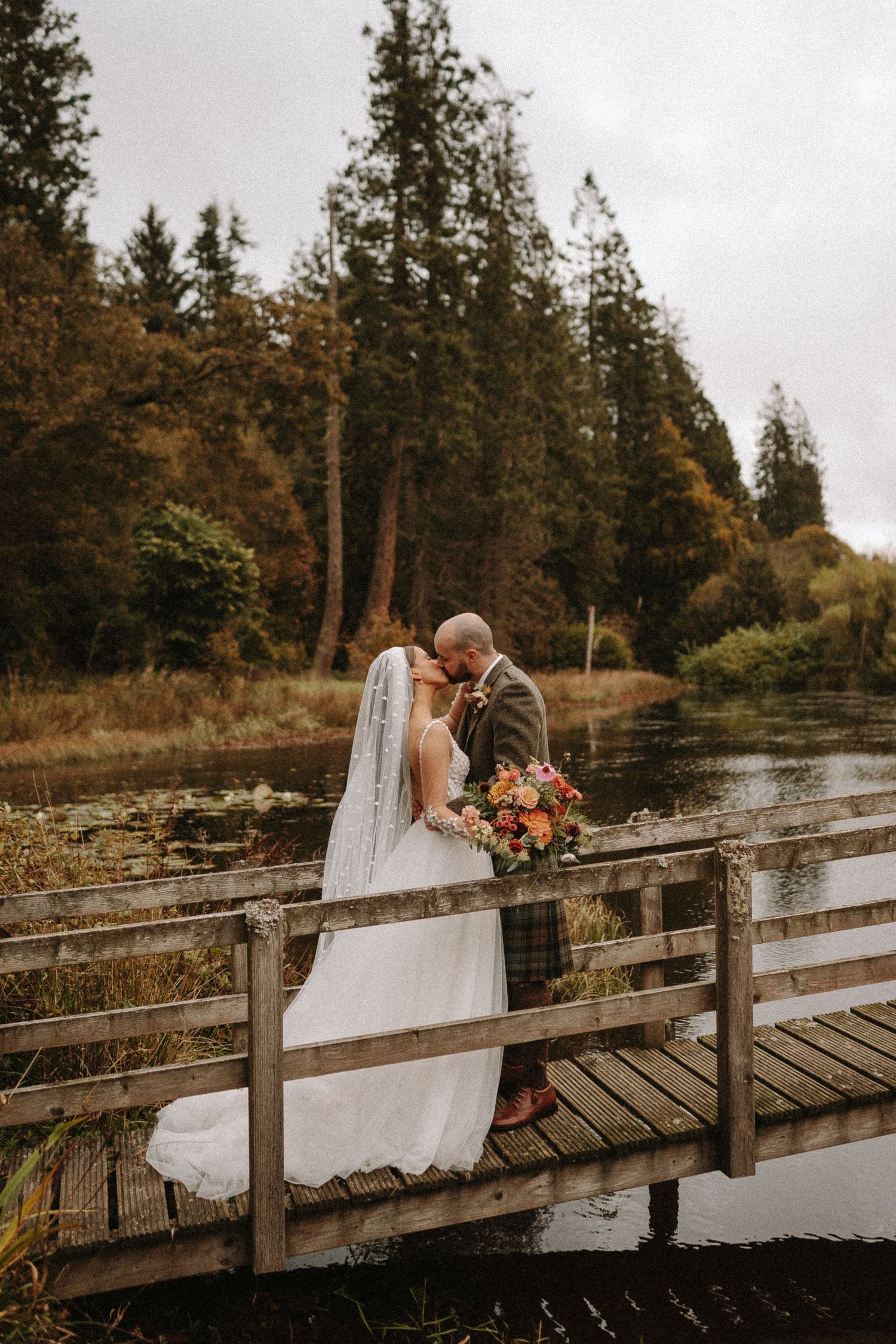 Wedding couple embracing on wooden bridge over pond with autumn forest backdrop. Aberdeen wedding photographer,  drumtochty castle
