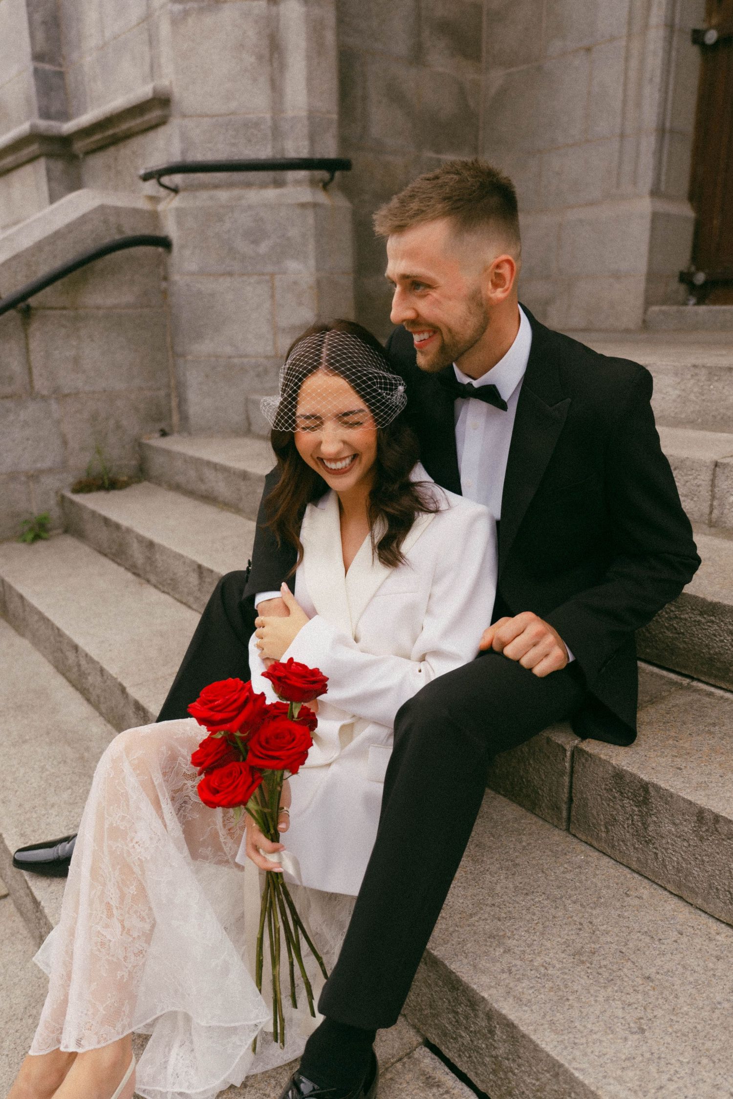 Two people share a candid laugh while sitting on stone steps in formal attire with red roses.