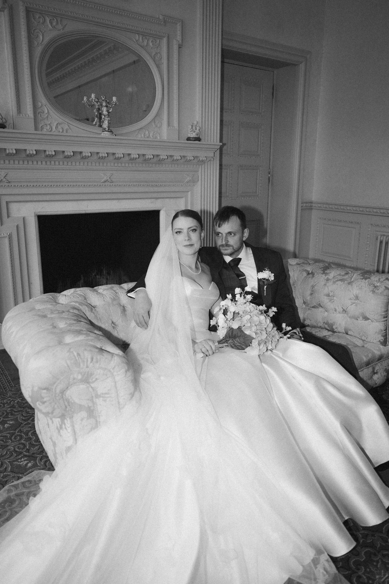 A newlywed couple sits together on an elegant sofa in front of an ornate fireplace. Mar Lodge Estate Wedding