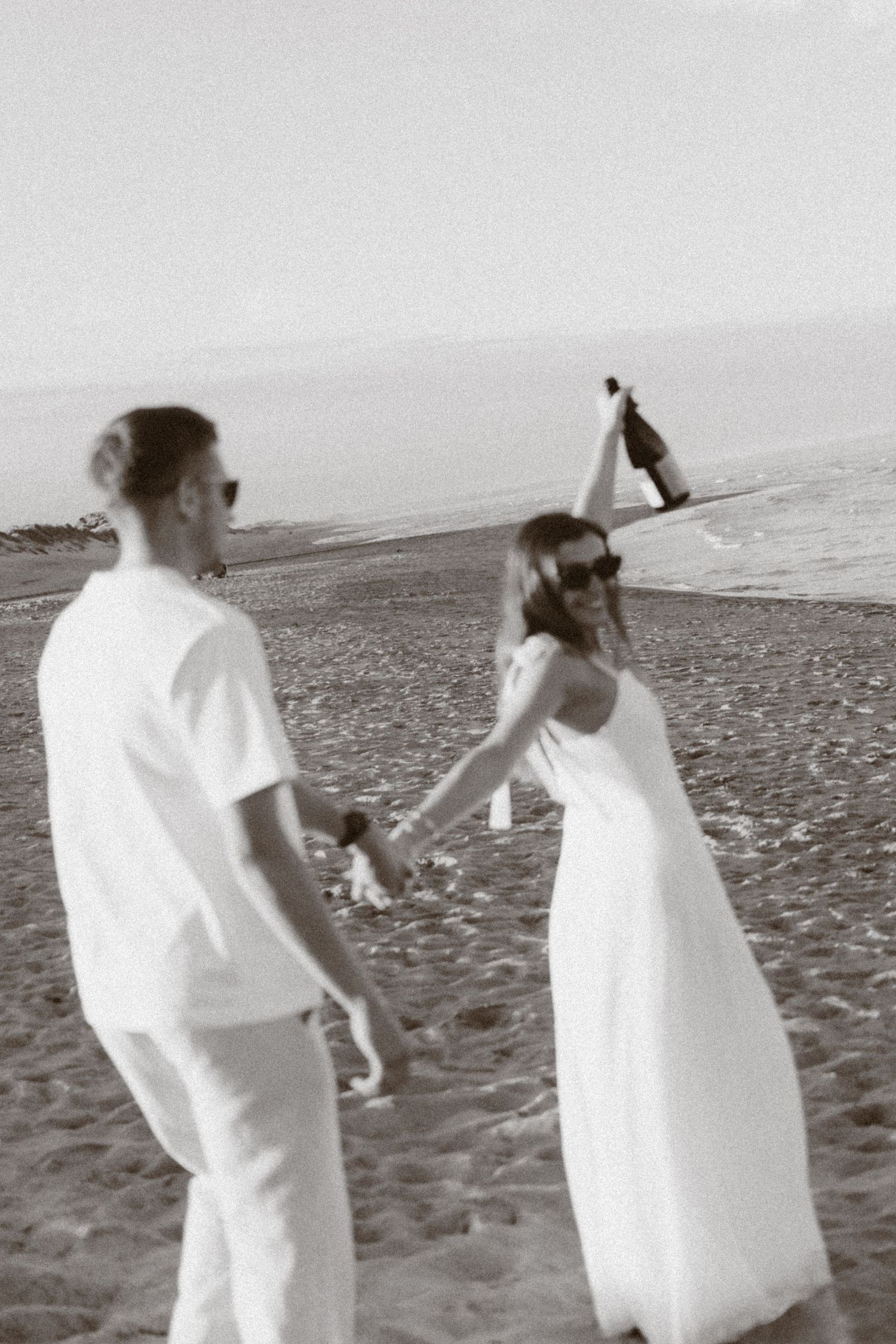 Black and white photo of a couple in white outfits holding hands on a sandy beach, woman holding a bottle.
