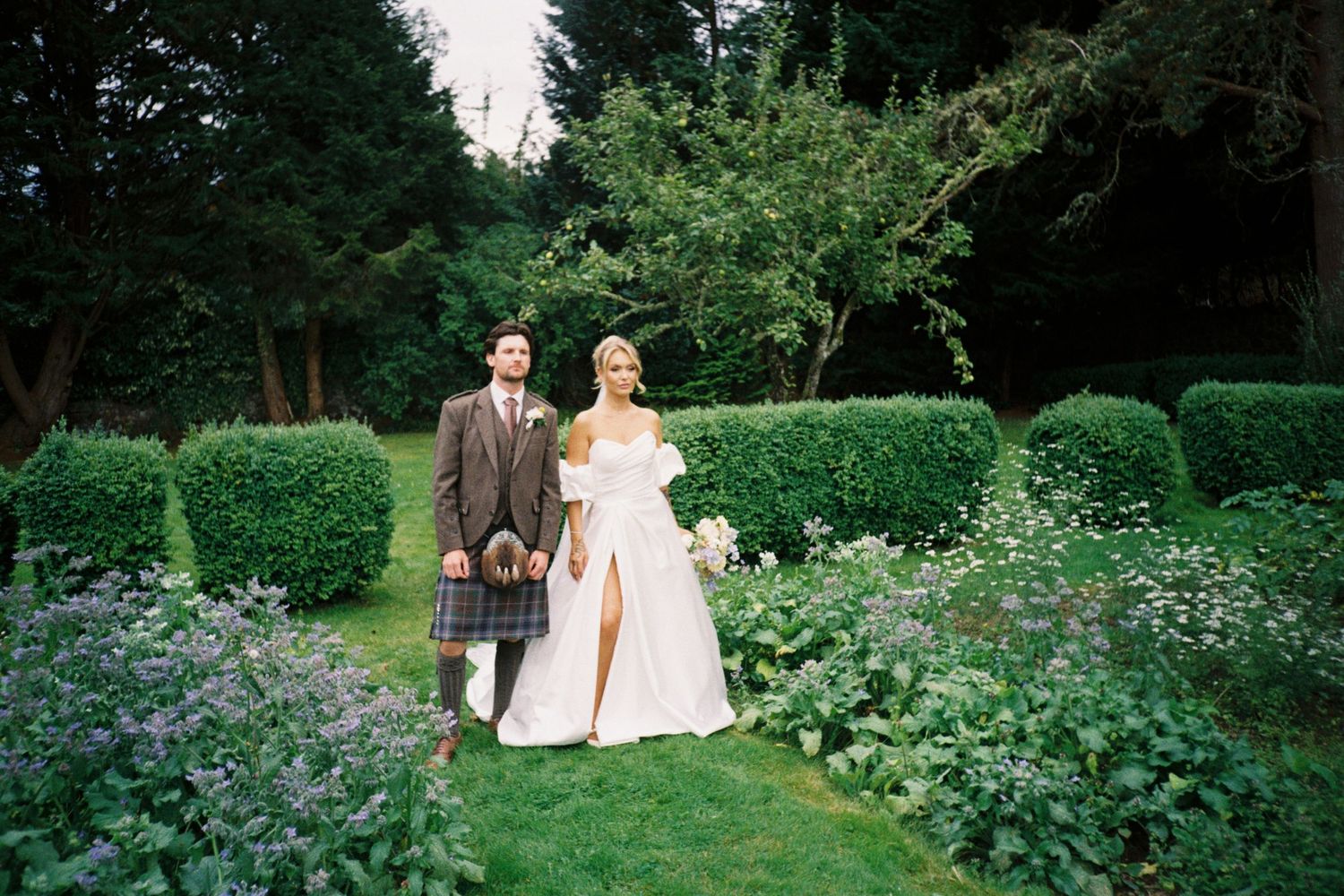 A couple walks through a lush garden path surrounded by green hedges and flowering plants on a summer day.