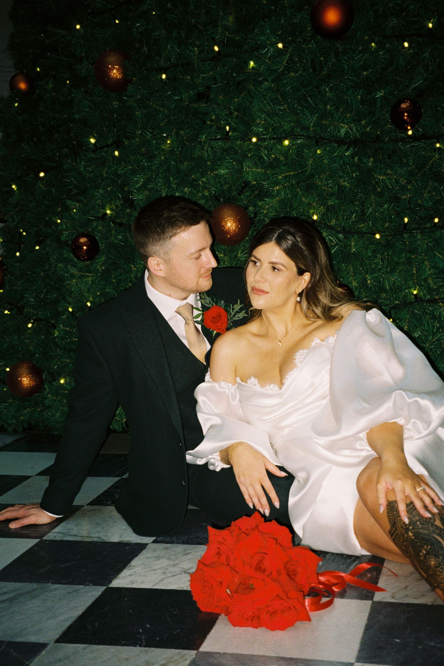A couple in formal attire shares an intimate moment on a checkered floor with red flowers and holiday decor in background.