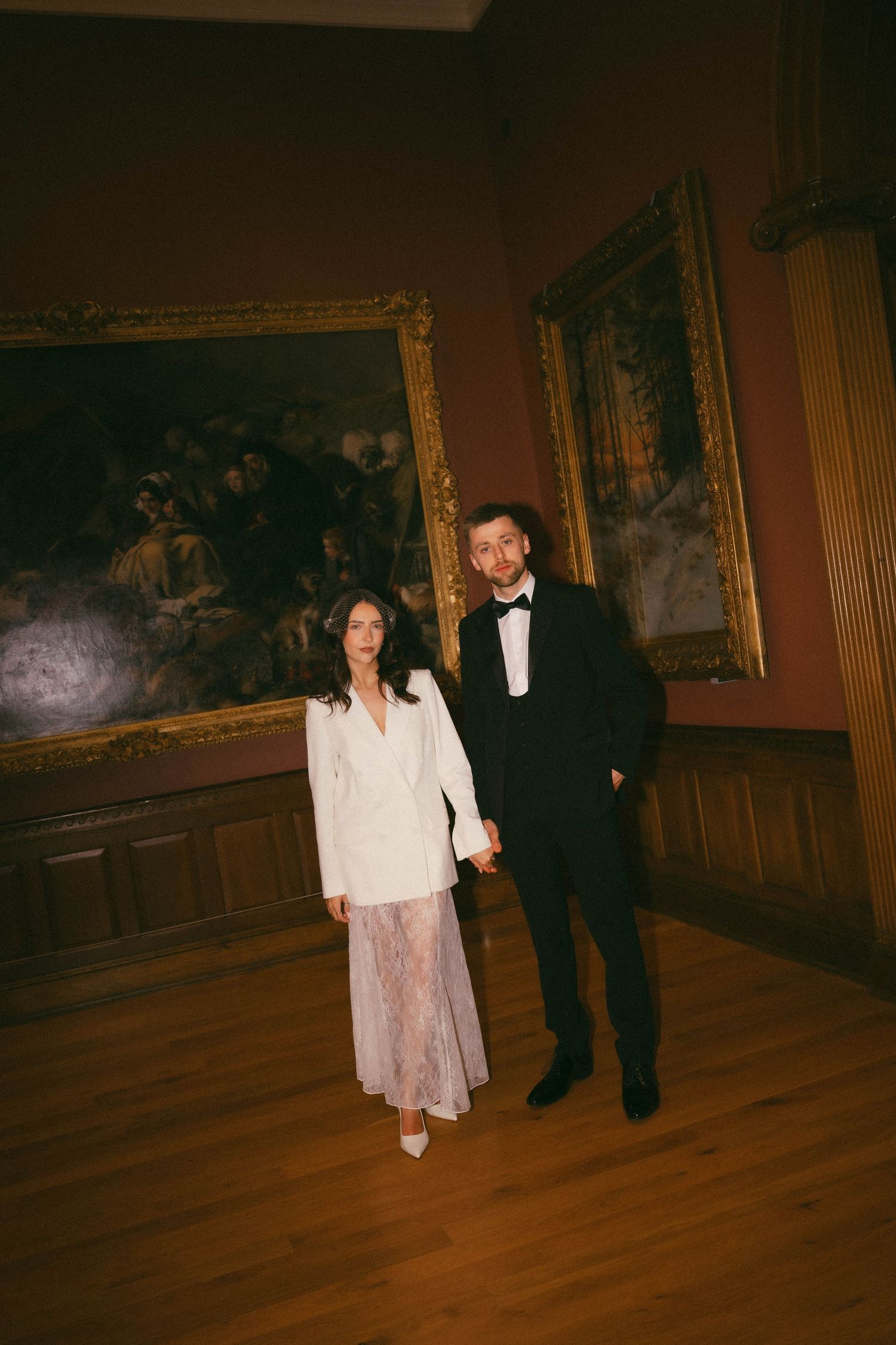 Couple in formal attire walks through an elegant museum gallery with wood floors and ornate paintings on walls.