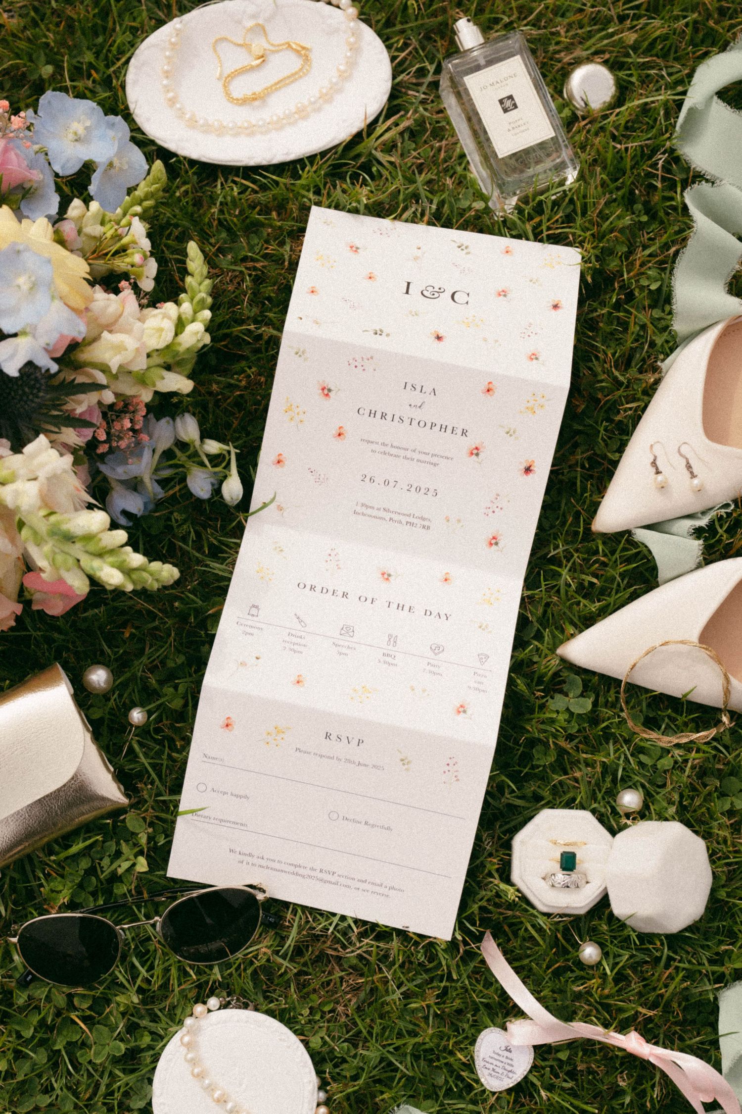 Wedding invitation on grass surrounded by flowers, perfume, shoes and decorative details in white and pink tones.
