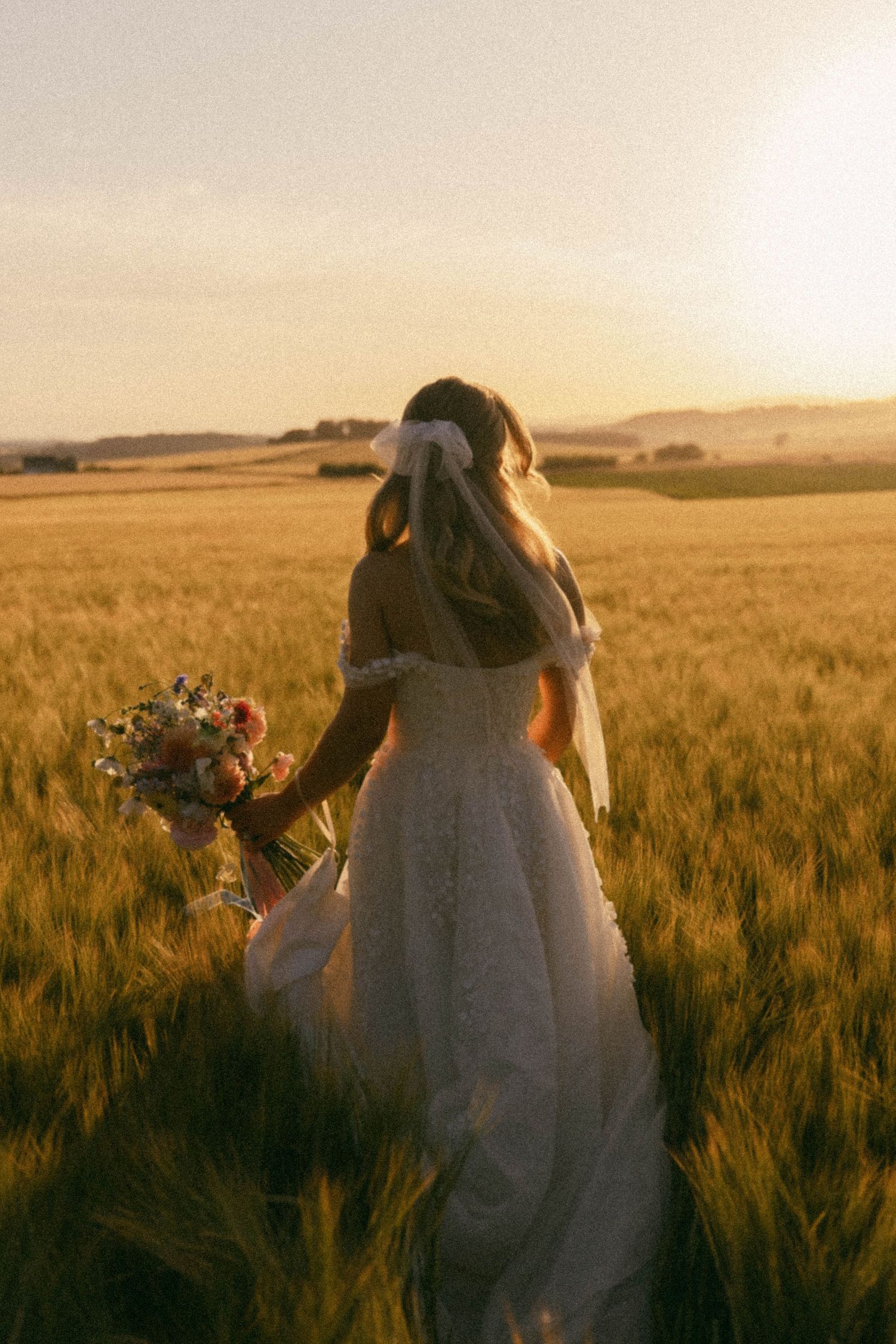 A figure in a white dress stands in a golden wheat field at sunset with a floral bouquet.