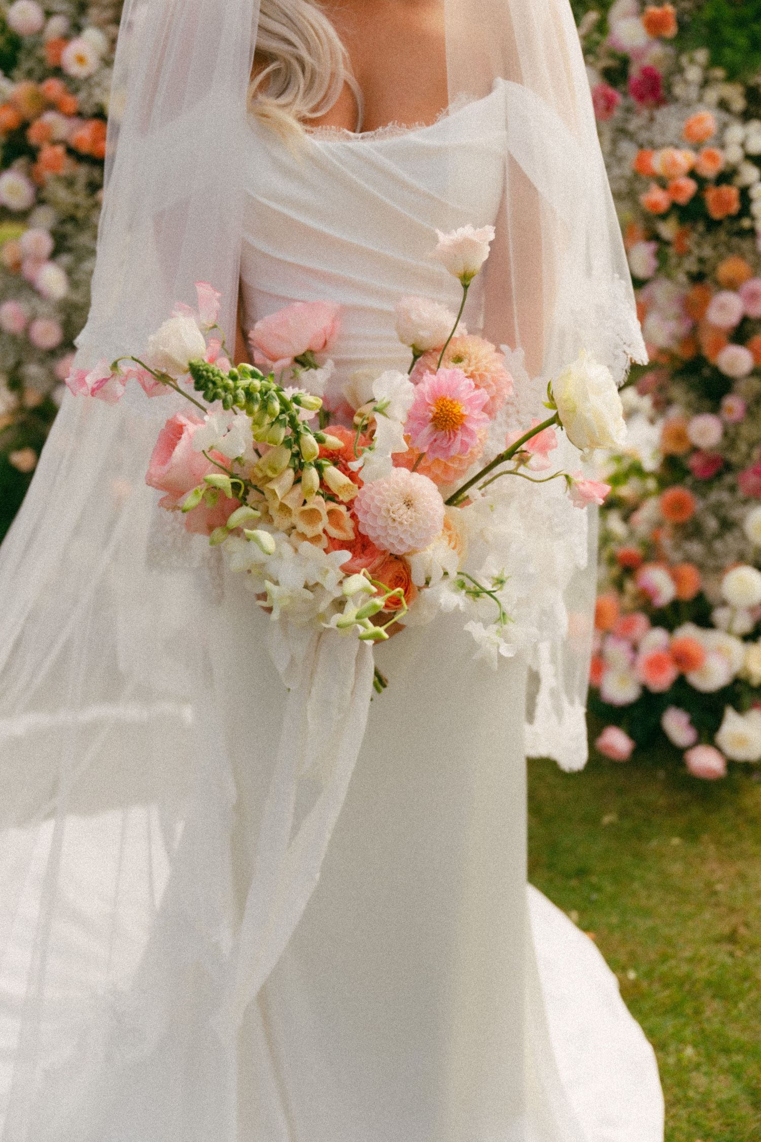 A wedding bouquet of pink and white flowers held against a flowing white dress with floral backdrop in garden setting.