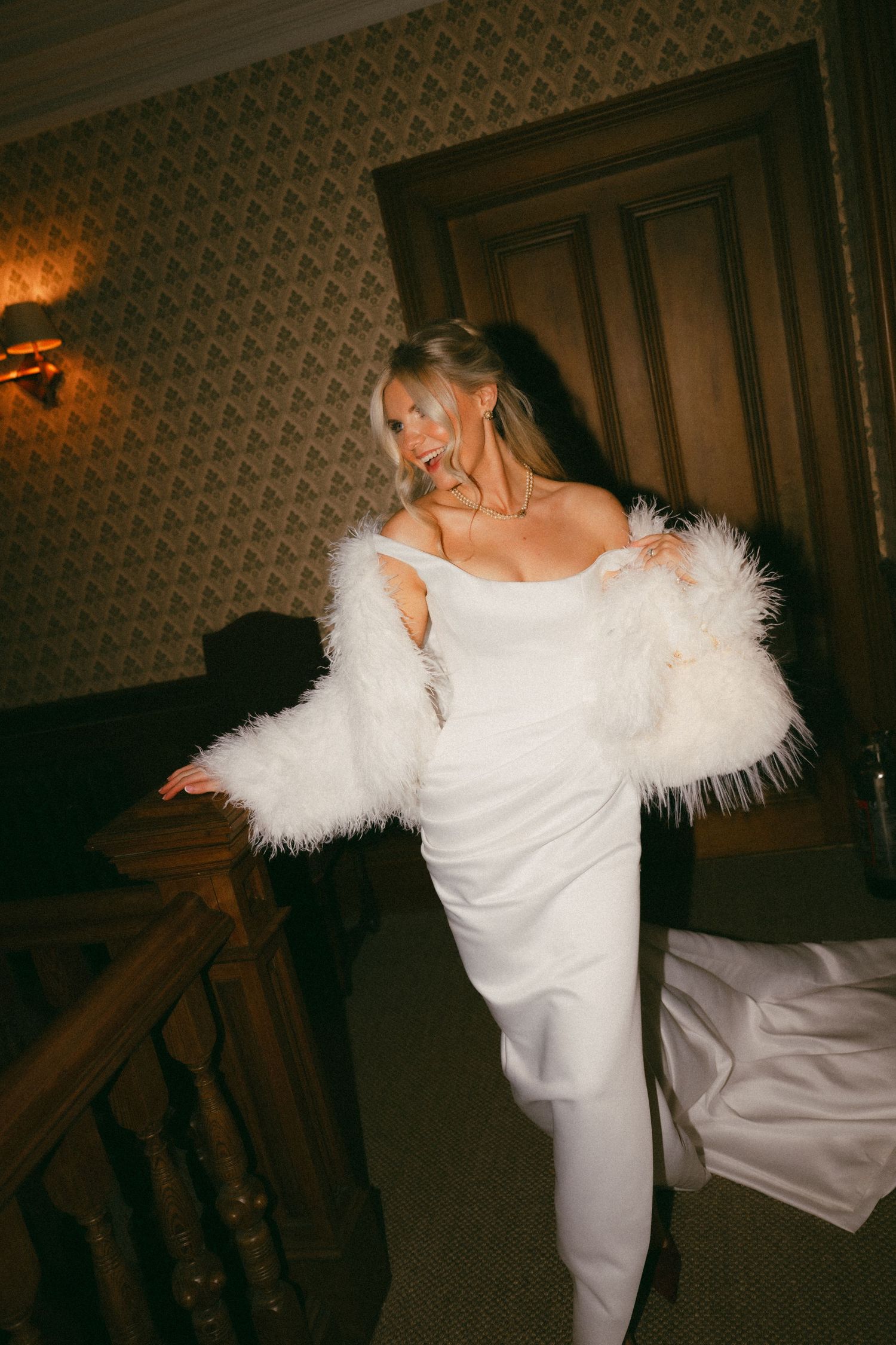 A person in an elegant white wedding gown with feathered sleeves poses on a staircase in warm lighting.