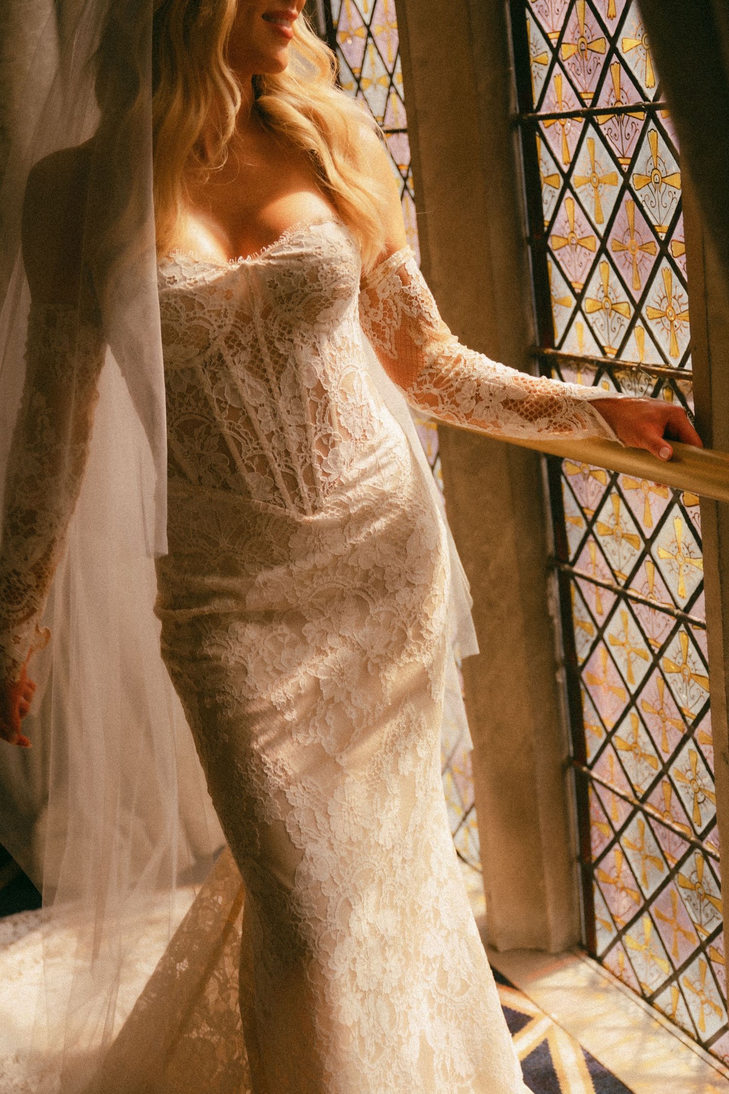 Bride in a fitted lace wedding gown with long sleeves and veil stands by a stained glass window in warm light.