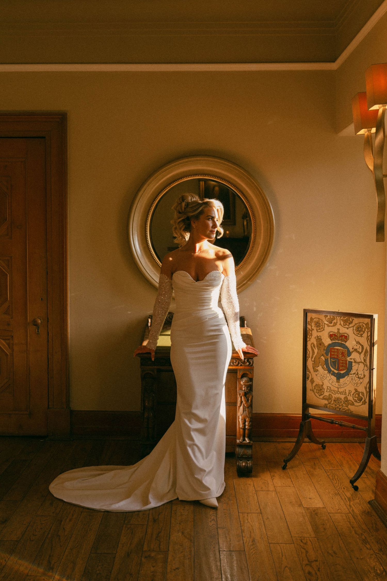 A bride in an elegant white off-shoulder gown with a long train poses in front of a circular mirror in a room with wood floors.