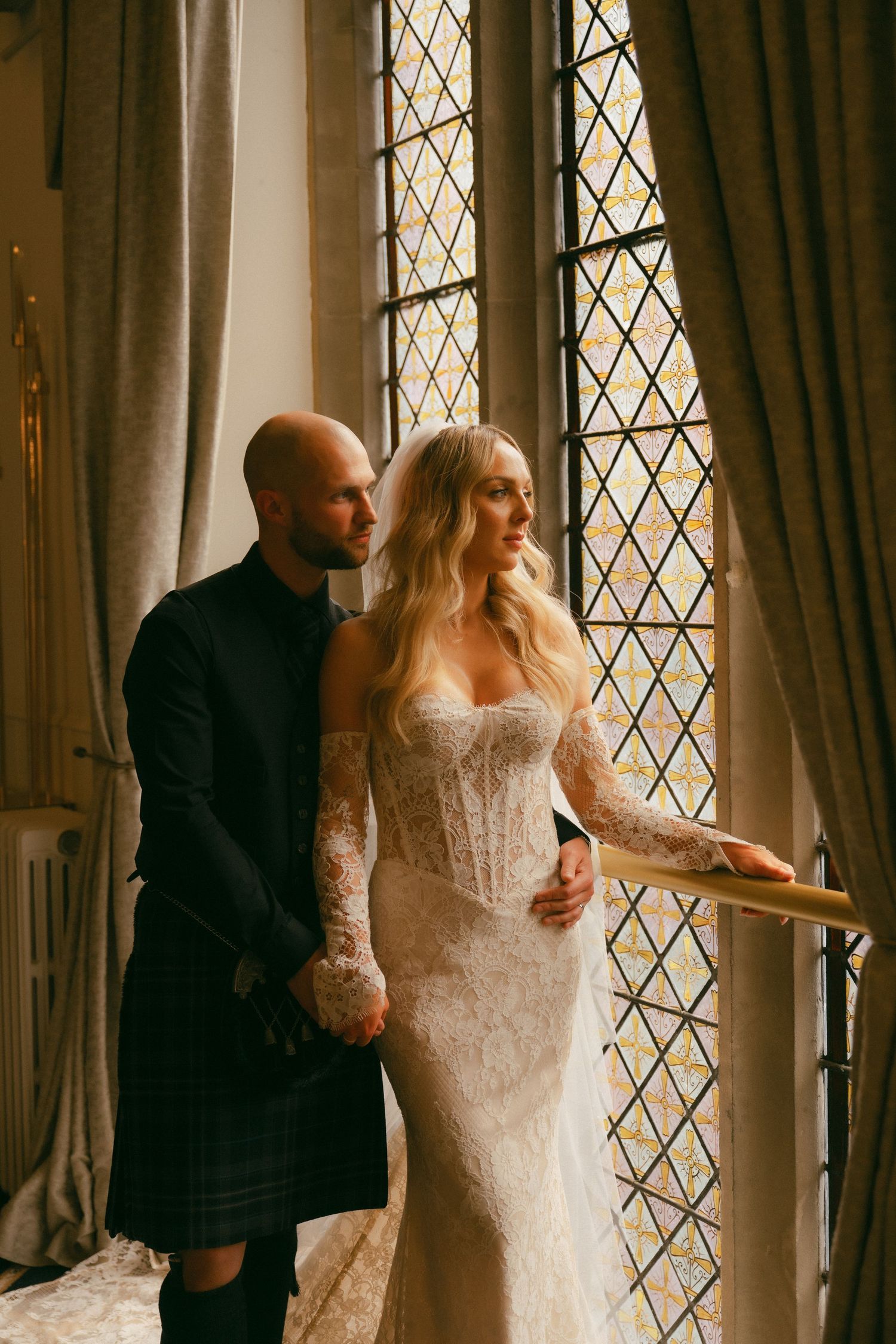 A bride in lace gown and groom in Scottish kilt stand together by a stained glass window in a castle.