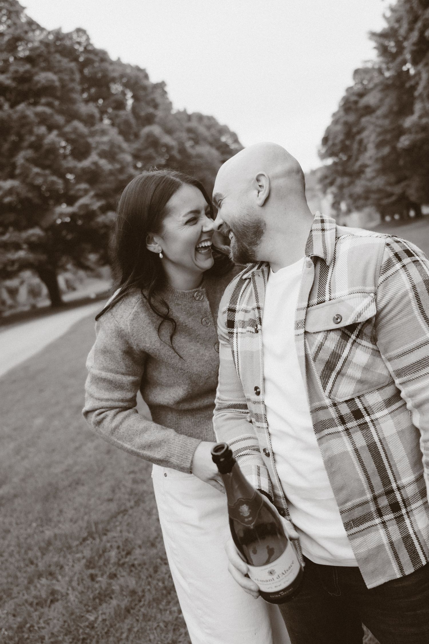 Black and white photo of a laughing couple sharing a romantic moment outdoors, holding a champagne bottle.