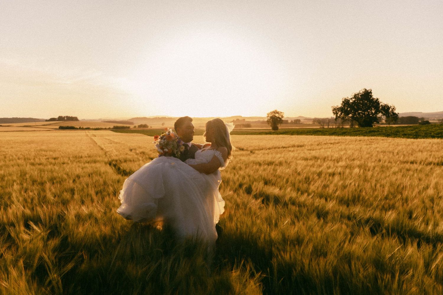 A couple in formal attire embrace in a golden wheat field at sunset creating a romantic silhouette against the warm sky.