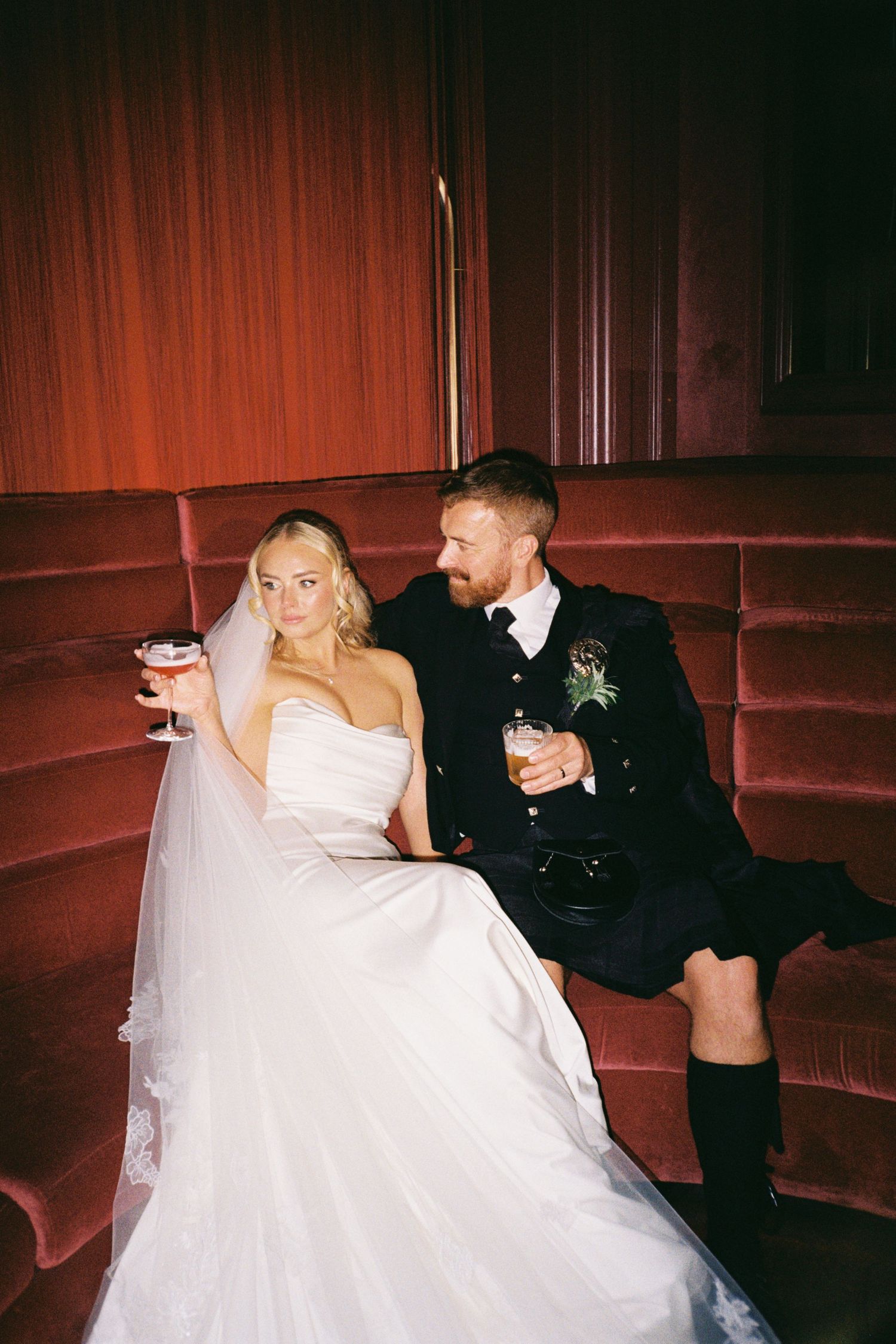 A couple in wedding attire relaxes on red velvet seating with drinks at a dimly lit venue.