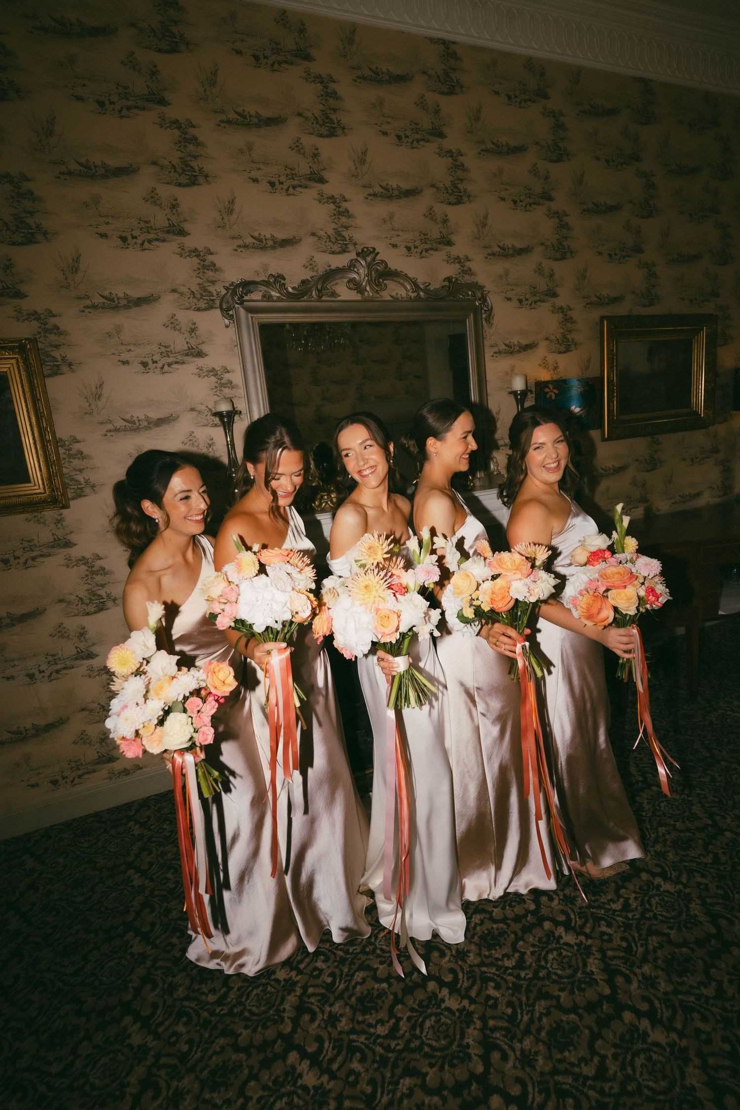 Bridesmaids in light-colored dresses laugh and smile while holding colorful pink and peach floral bouquets.