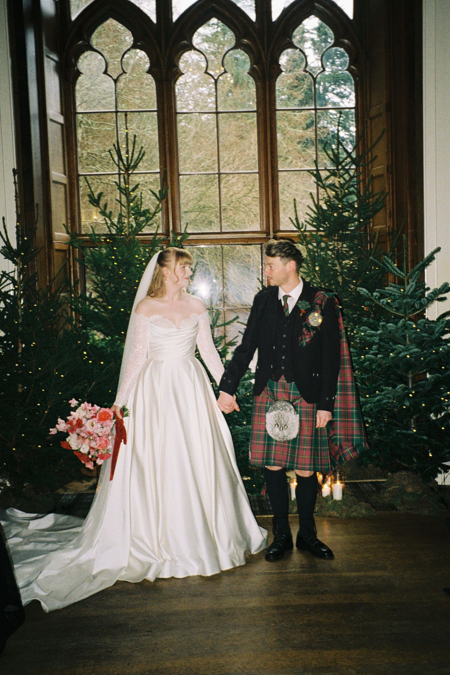 Wedding couple poses in front of gothic windows with Christmas trees and traditional Scottish kilts in elegant venue.