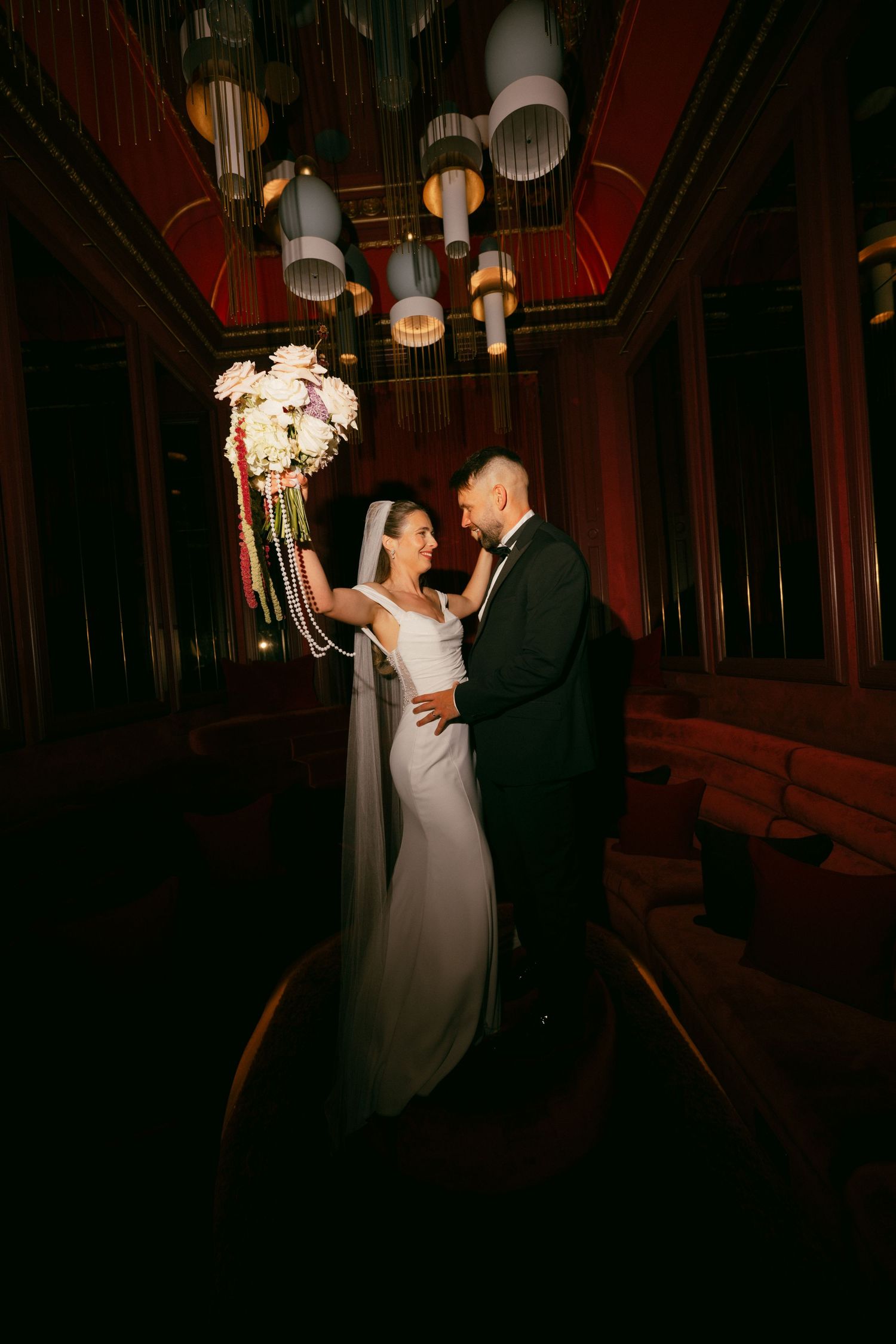 Bride and groom share romantic moment in dimly lit venue, bride holding white bouquet aloft while wearing veil and gown.