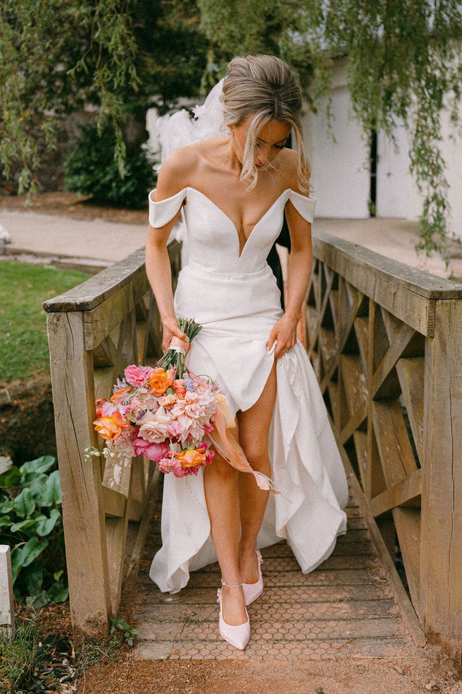 Bride in off-shoulder white gown holding colorful bouquet while crossing wooden bridge.  Aberdeen wedding photographer, douneside house