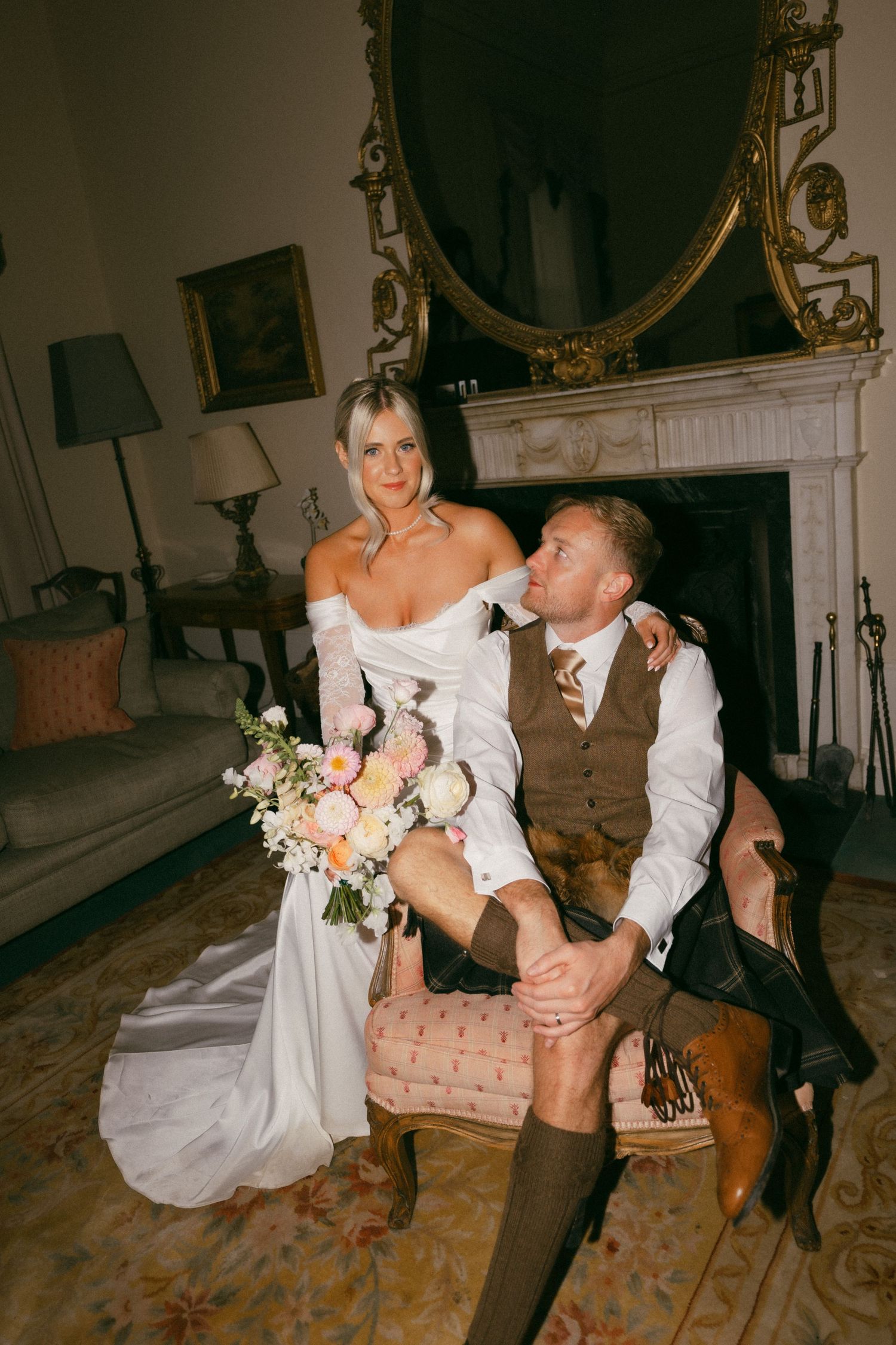 Couple in wedding attire sits on vintage pink chair by ornate fireplace with flowers and elegant decor.