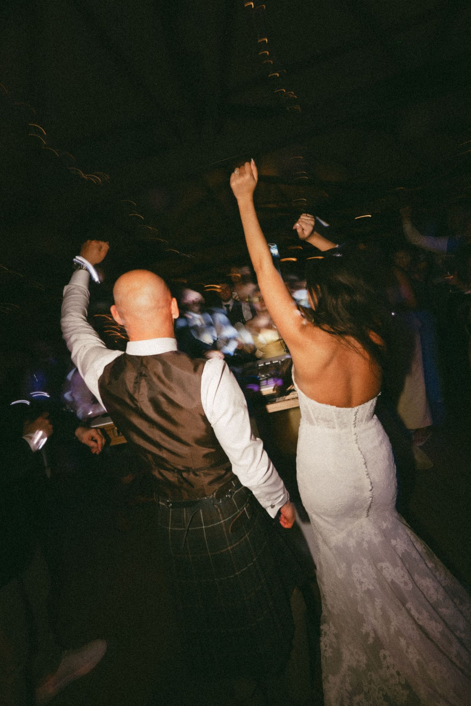 Bride and groom dancing joyfully at wedding reception, groom wearing kilt, bride in backless lace gown, arms raised.