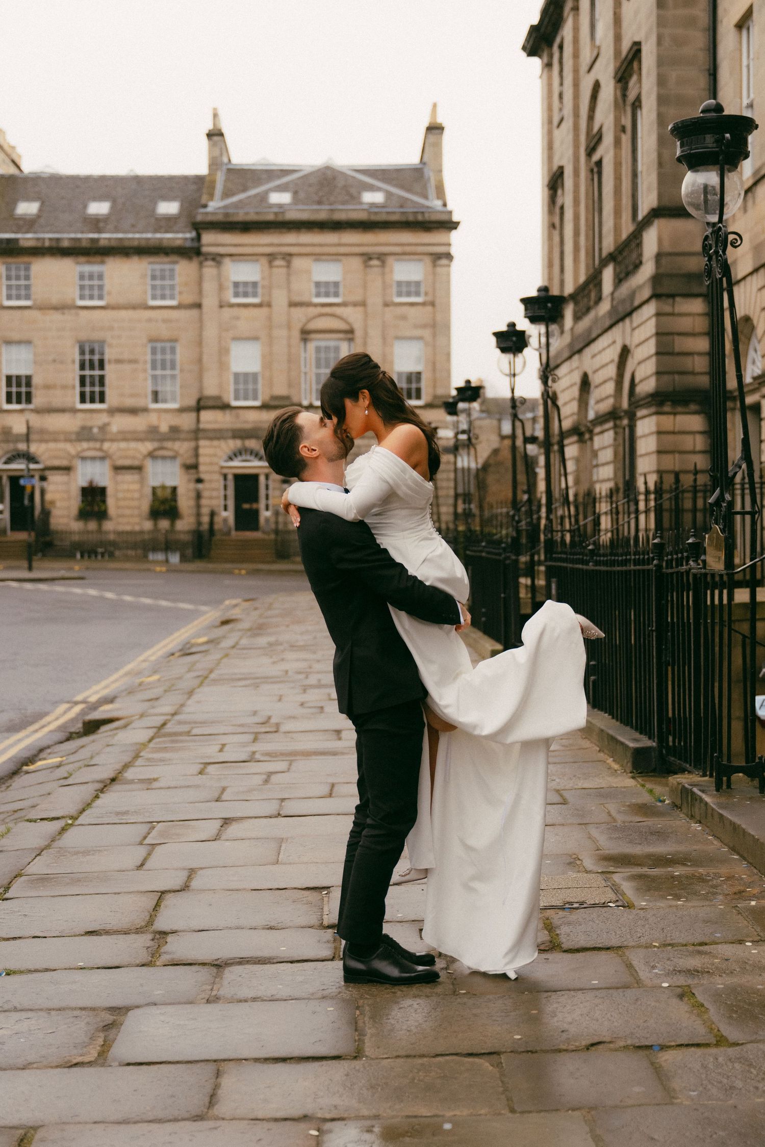 A couple embraces on a cobblestone street in Bath, England with historic Georgian architecture in the background.