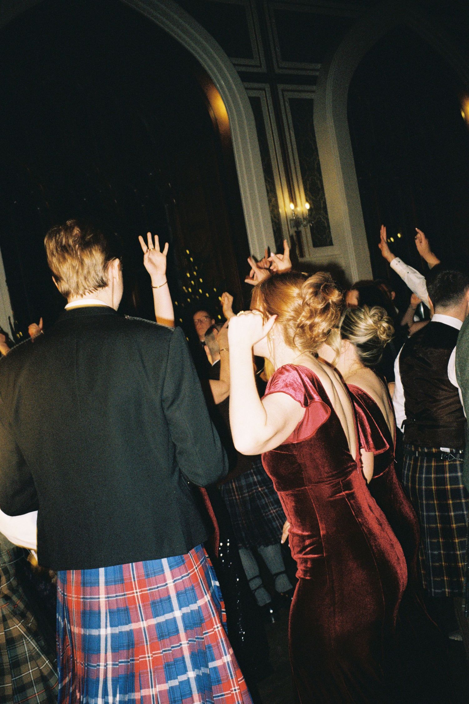 People dancing at a formal event wearing traditional Scottish kilts and formal attire in a dimly lit venue.