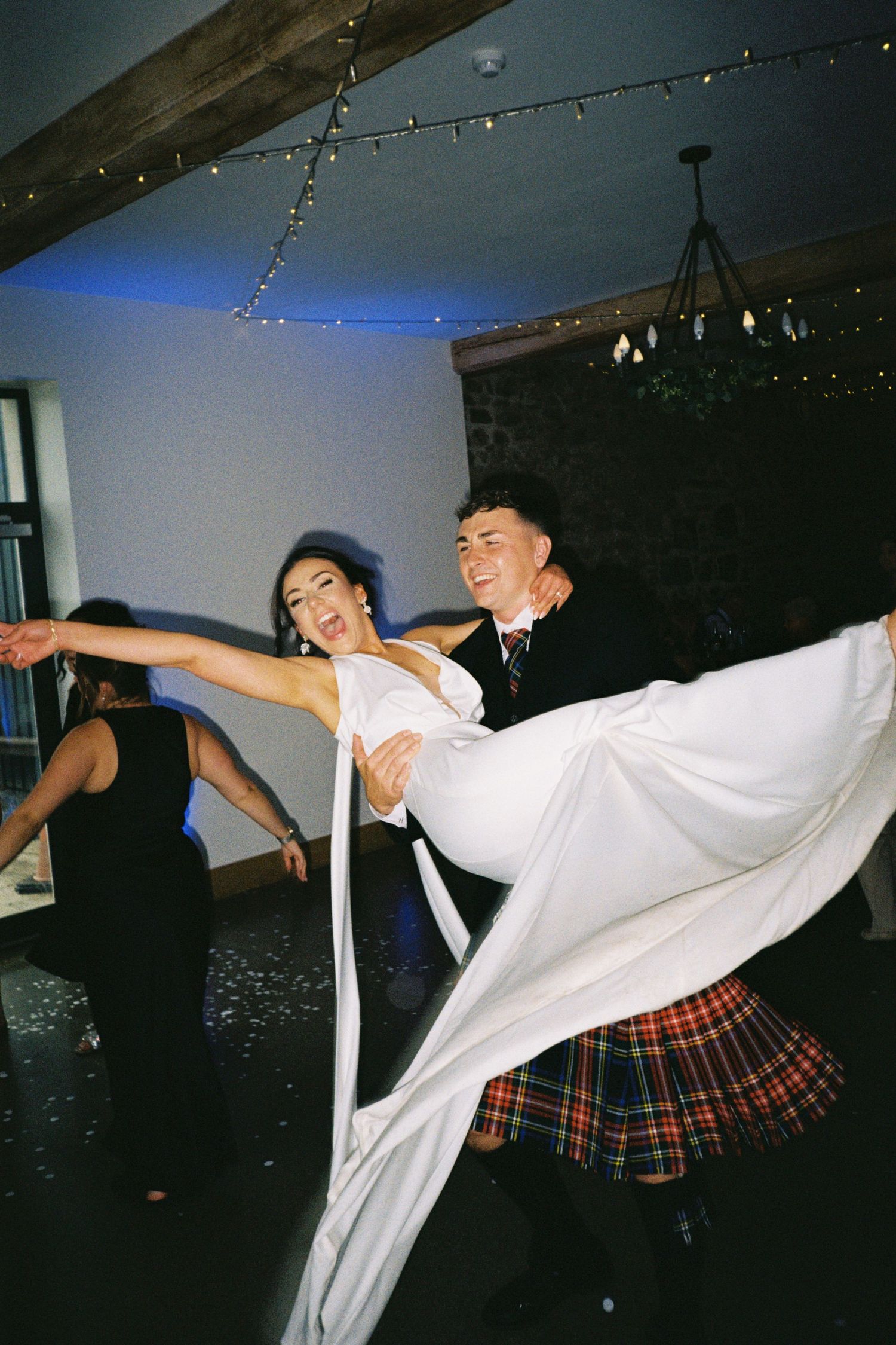Two dancers in white shirts and plaid skirts perform energetic dance moves under string lights in a dark room.