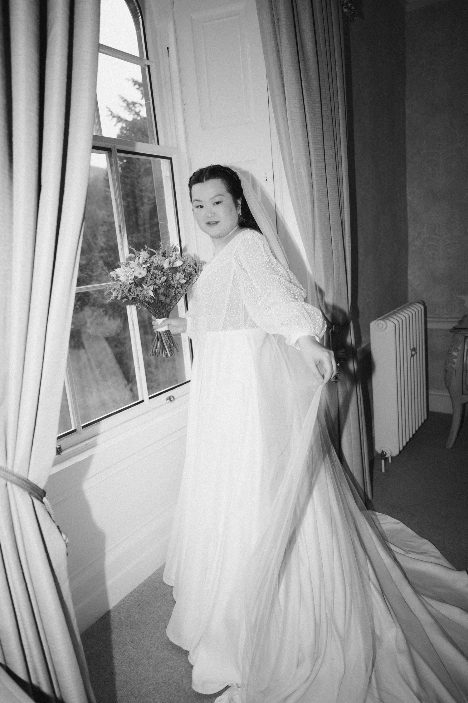 A bride in a flowing white dress stands by a window holding a bouquet in black and white photography. Drumtochty Castle