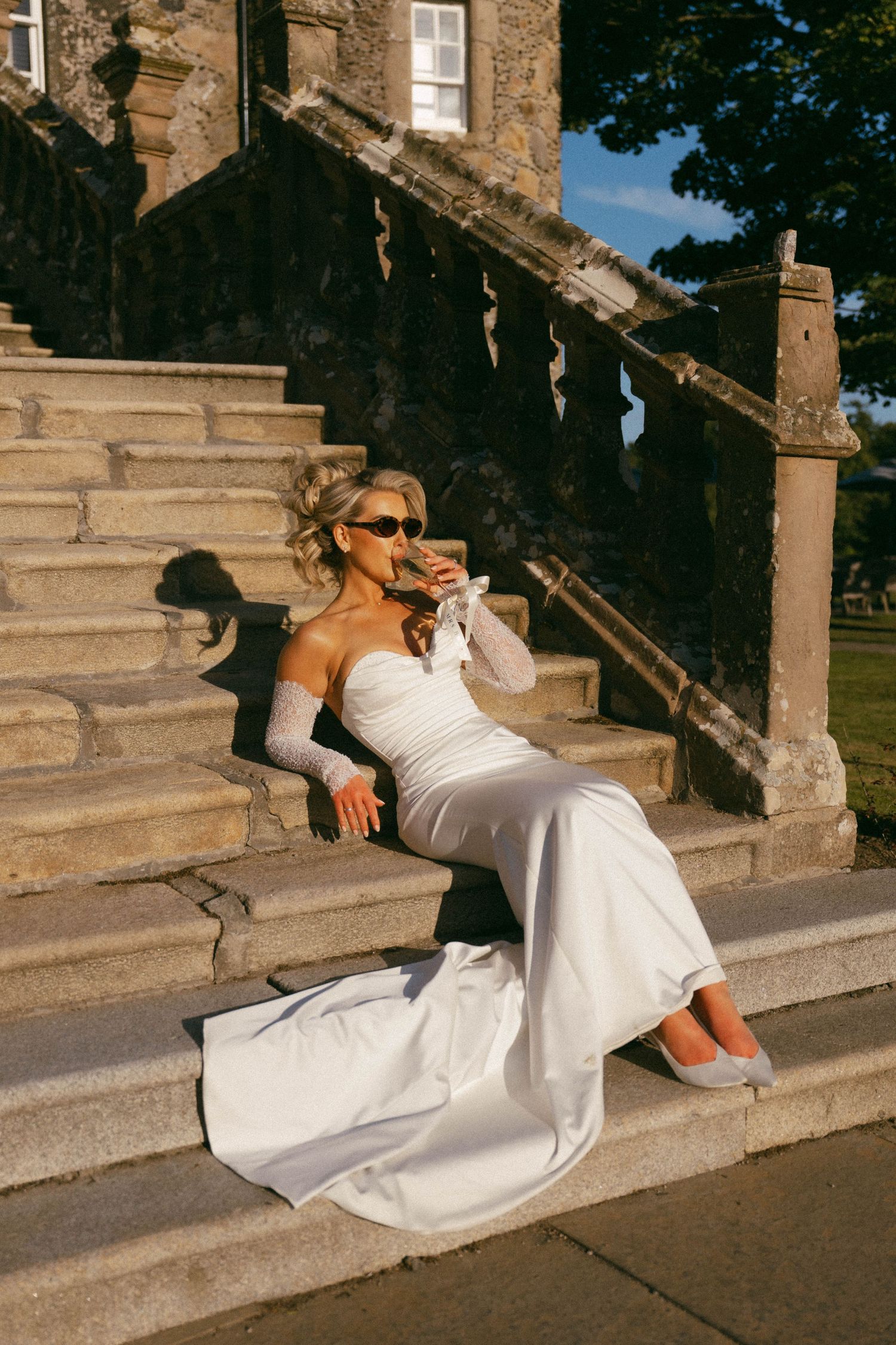 Bride in white strapless gown relaxing on stone steps while wearing sunglasses on sunny day.