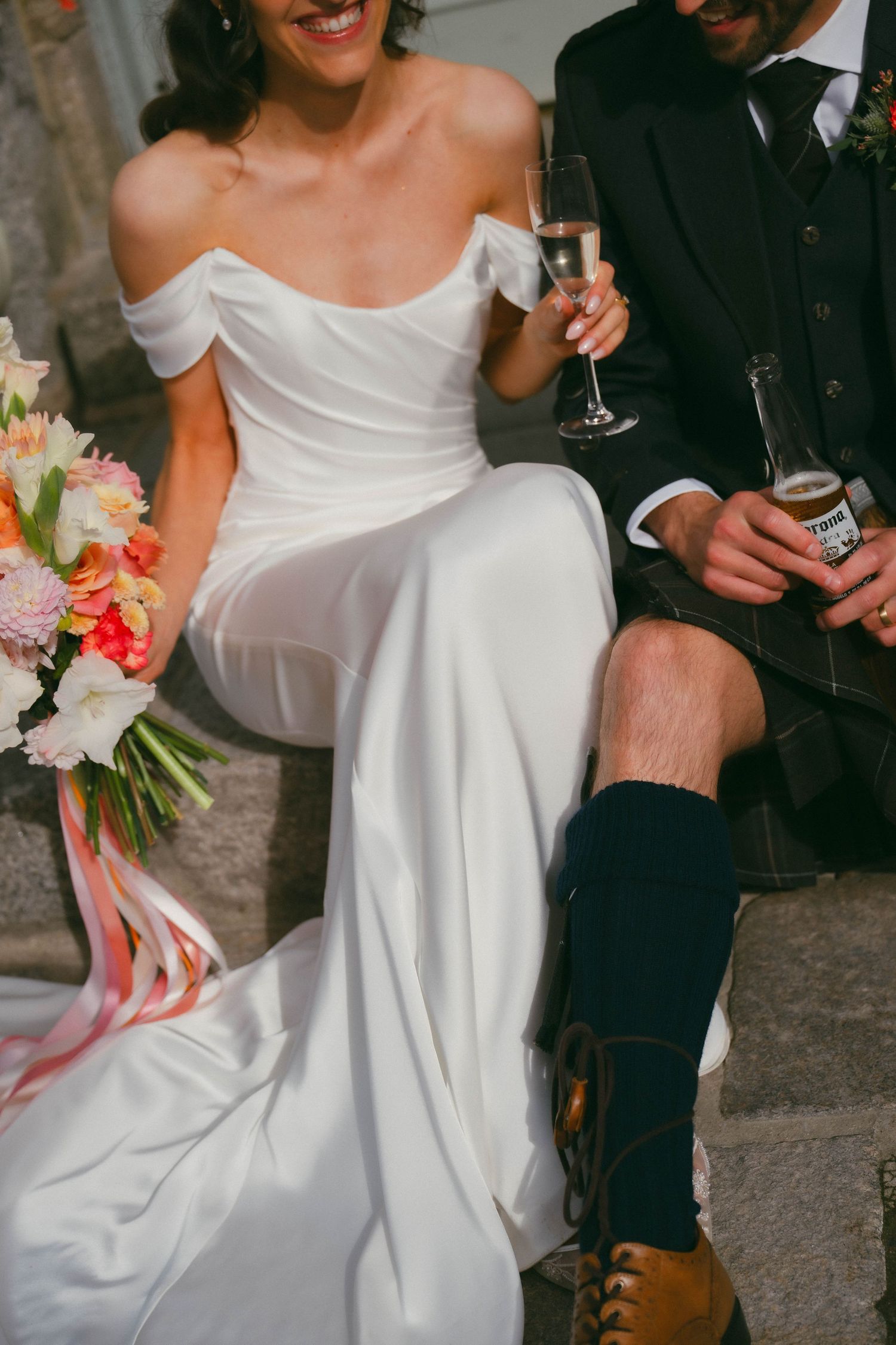 A wedding couple poses while holding champagne glasses, with the bride wearing a flowing white dress against a stone wall.